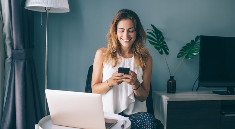 Influencerin sitzt mit Smartphone und Laptop in ihrem Wohnzimmer.