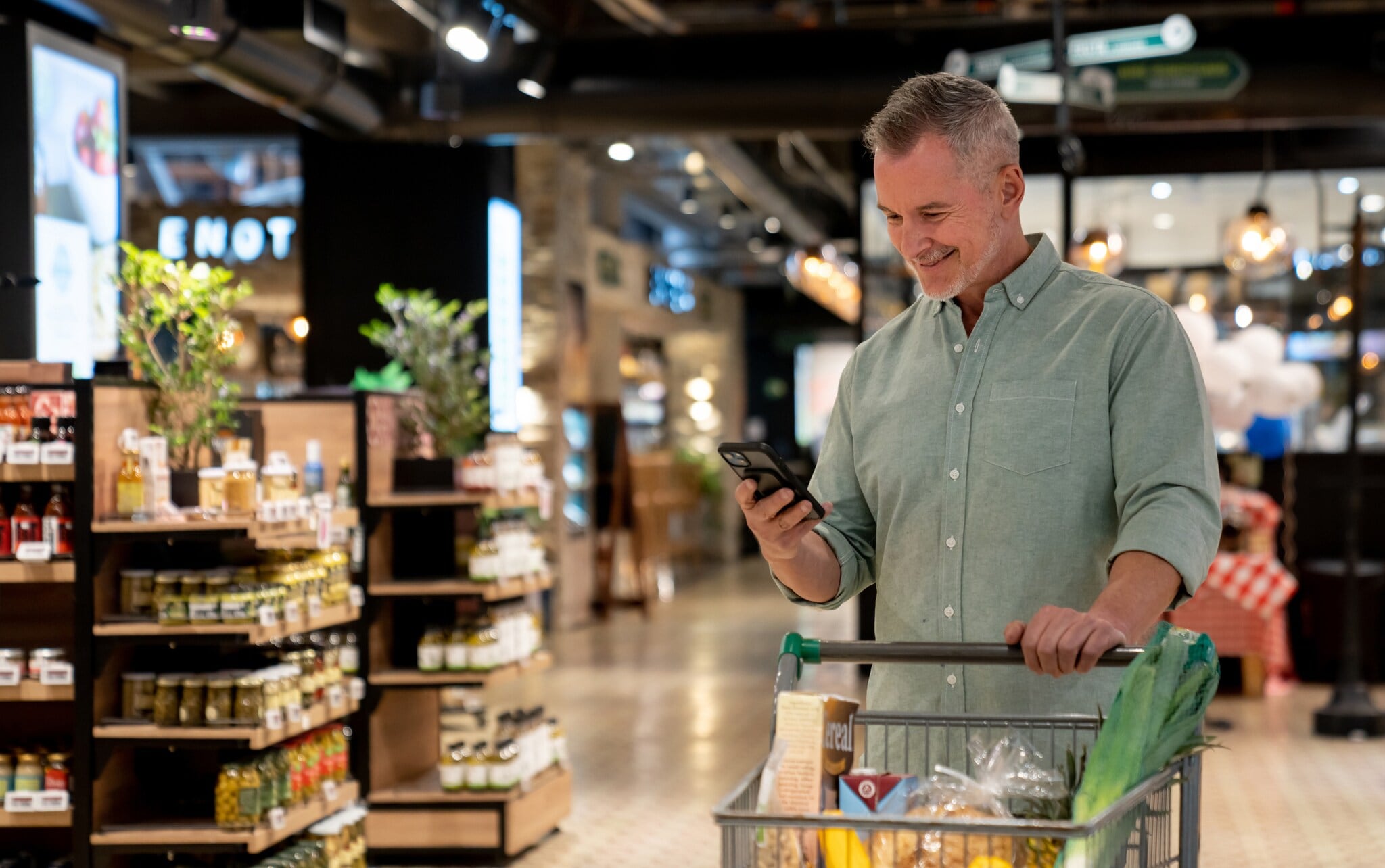 Eine Person steht mit einem Einkaufswagen im Supermarkt und schaut auf ihr Handy in der Hand. Eine Person steht mit einem Einkaufswagen im Supermarkt und schaut auf ihr Handy in der Hand.