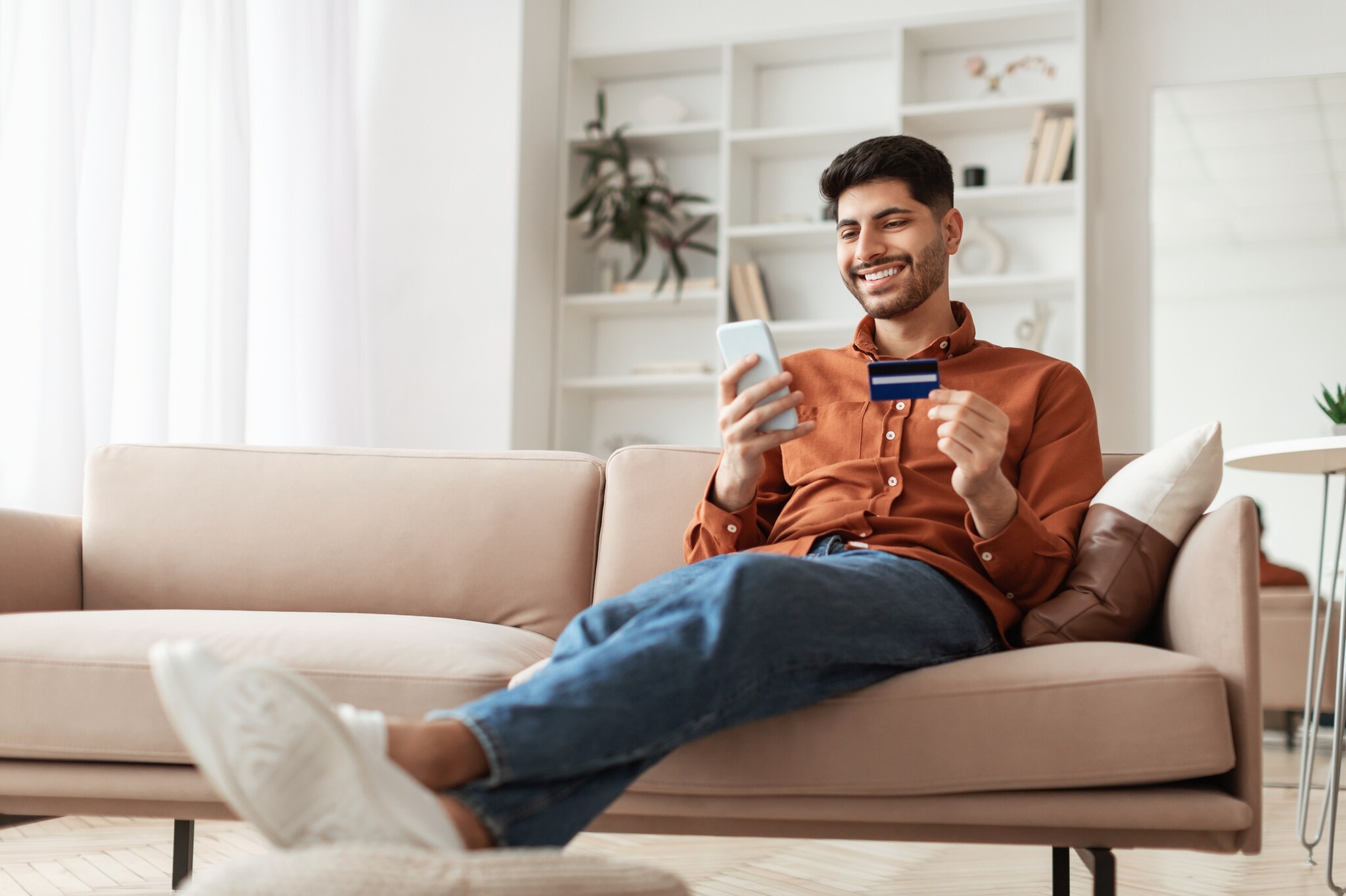Prostock-Studio_iStock_1368987043-1 Eine Person sitzt mit ausgestreckten Beinen auf einem Sofa und hält eine Kreditkarte sowie ein Smartphone in den Händen.