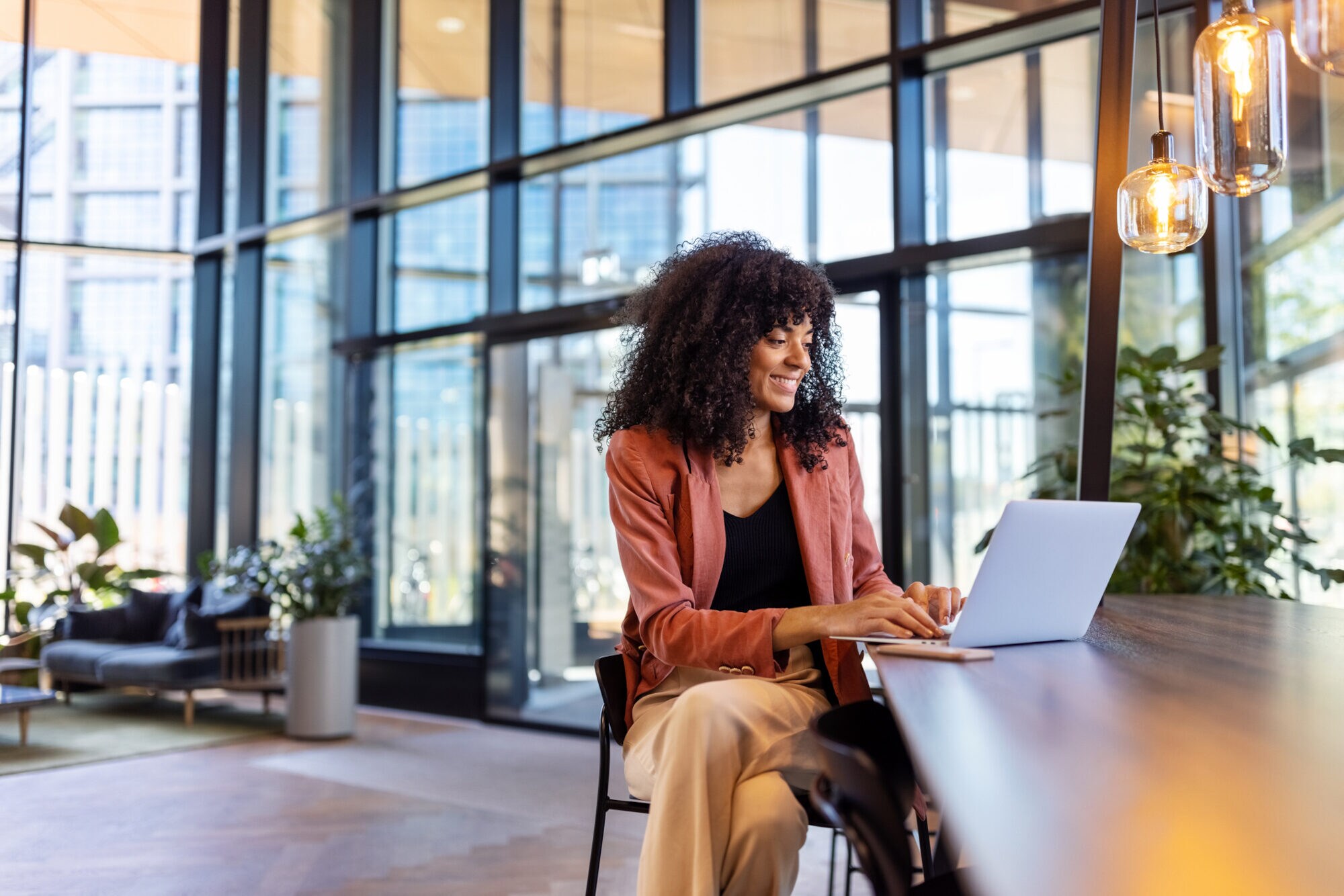 Eine Person sitzt in einem lichtdurchfluteten Ambiente an einem hochbeinigen Tisch und arbeitet am Laptop.
