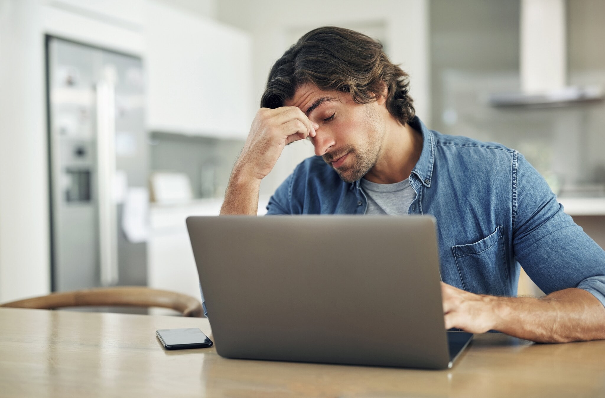 4361 Ein Mann sitzt vor einem Laptop an einem Tisch, hat die Augen geschlossen und eine Hand an seine Stirn gelegt.