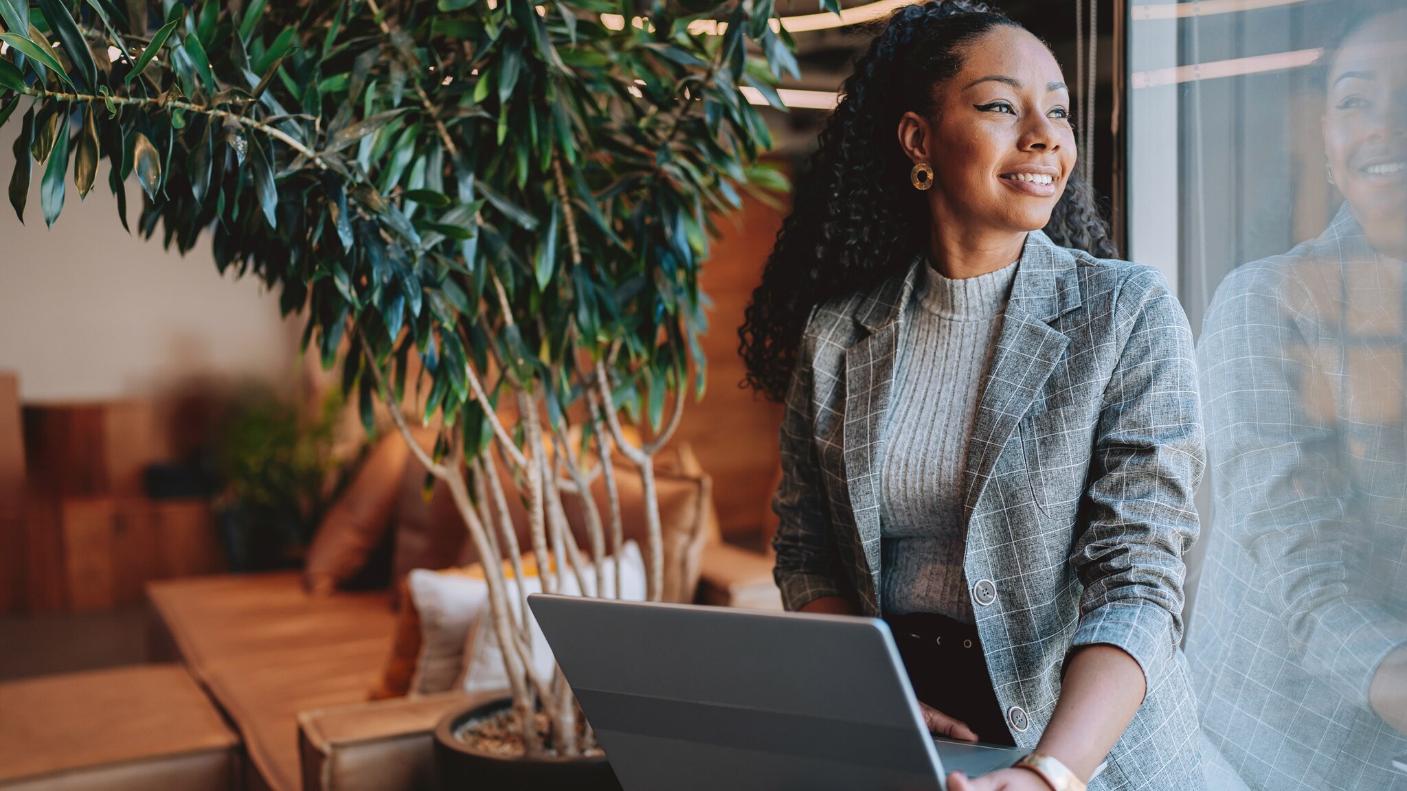 Eine Frau im grauen Blazer sitzt mit Laptop auf dem Schoß an einem Fenster in einer Lounge mit Topfpflanze und schaut zufrieden lächelnd hinaus.