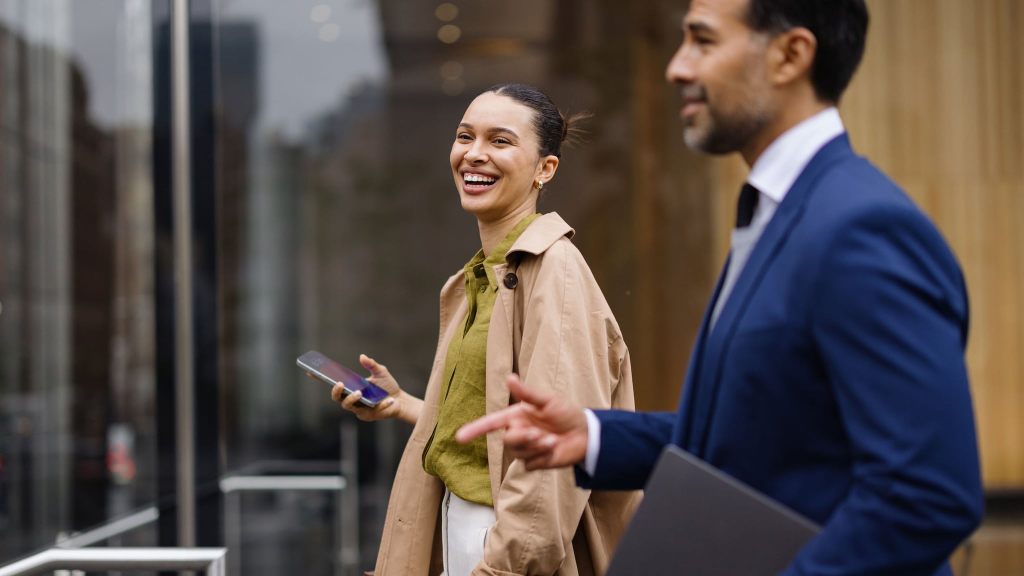 Eine gut gelaunte Frau mit Smartphone in der Hand und ein Mann im blauen Anzug und Laptop unter dem Arm gehen vor einem Bürogebäude nebeneinanderher.