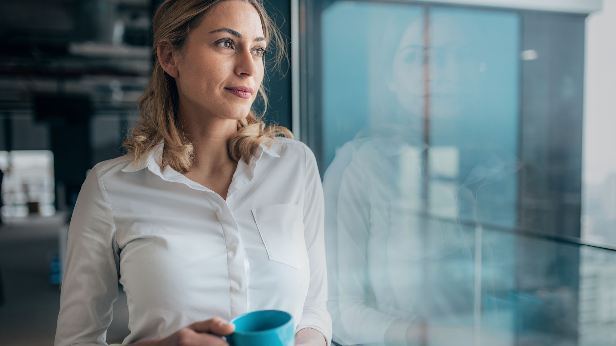 Eine Frau in weißer Bluse steht mit blauer Kaffeetasse in der Hand an einem Fenster in einem Büro.