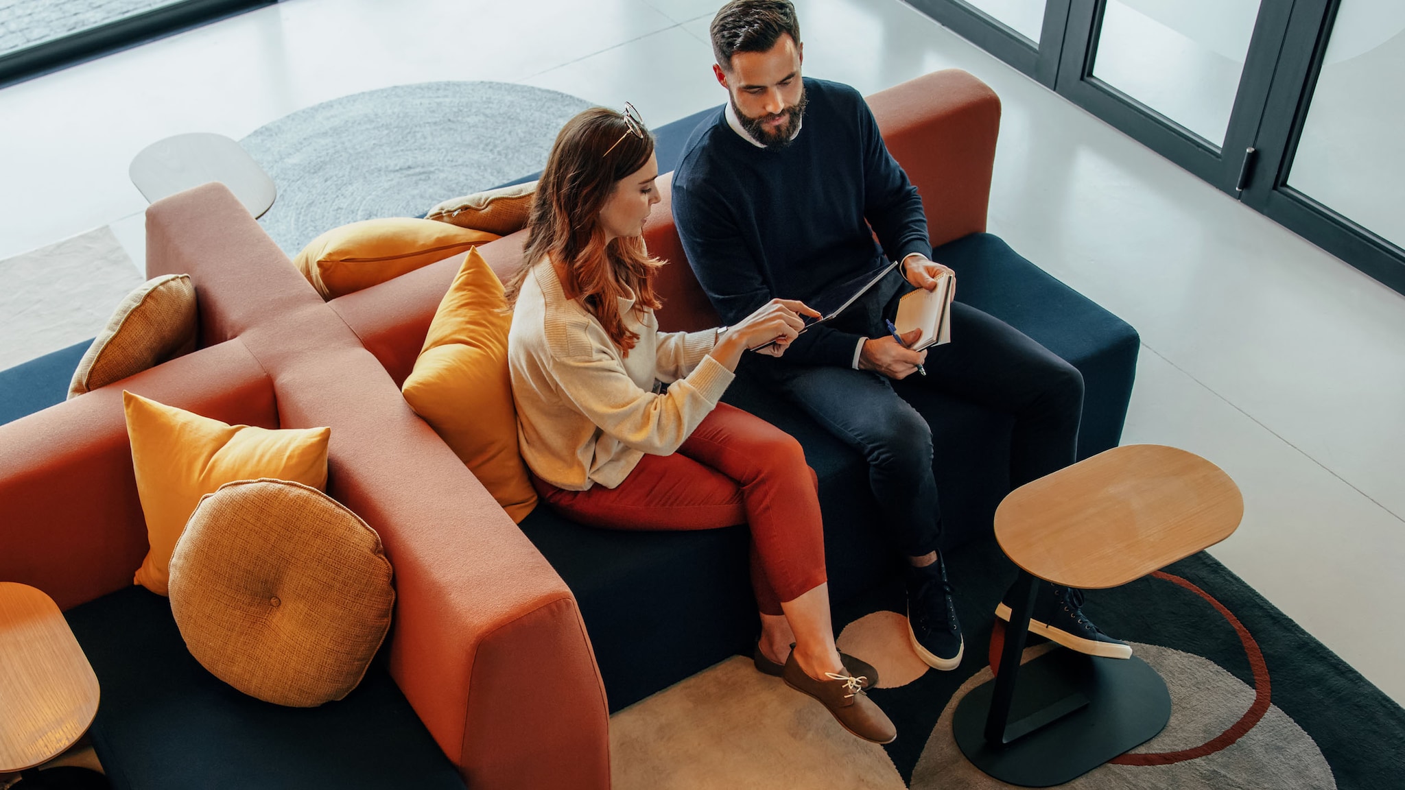 Eine Frau und ein Mann sitzen auf einer modularen Couchgarnitur und arbeiten gemeinsam mit einem Tablet in einer modernen Lobby.