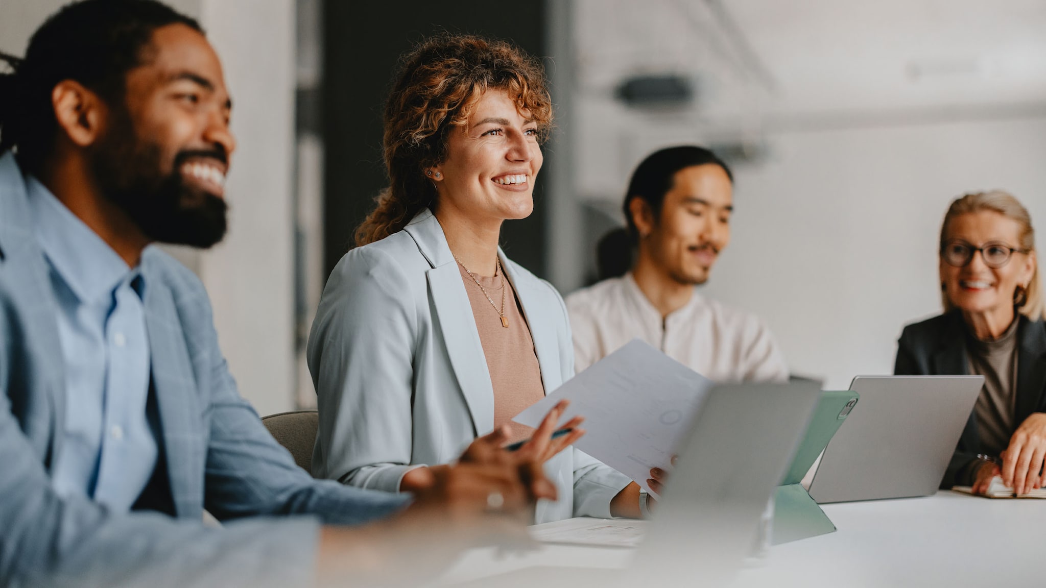 Vier lächelnde Personen sitzen nebeneinander an einem Konferenztisch mit Laptops in einem Büro.