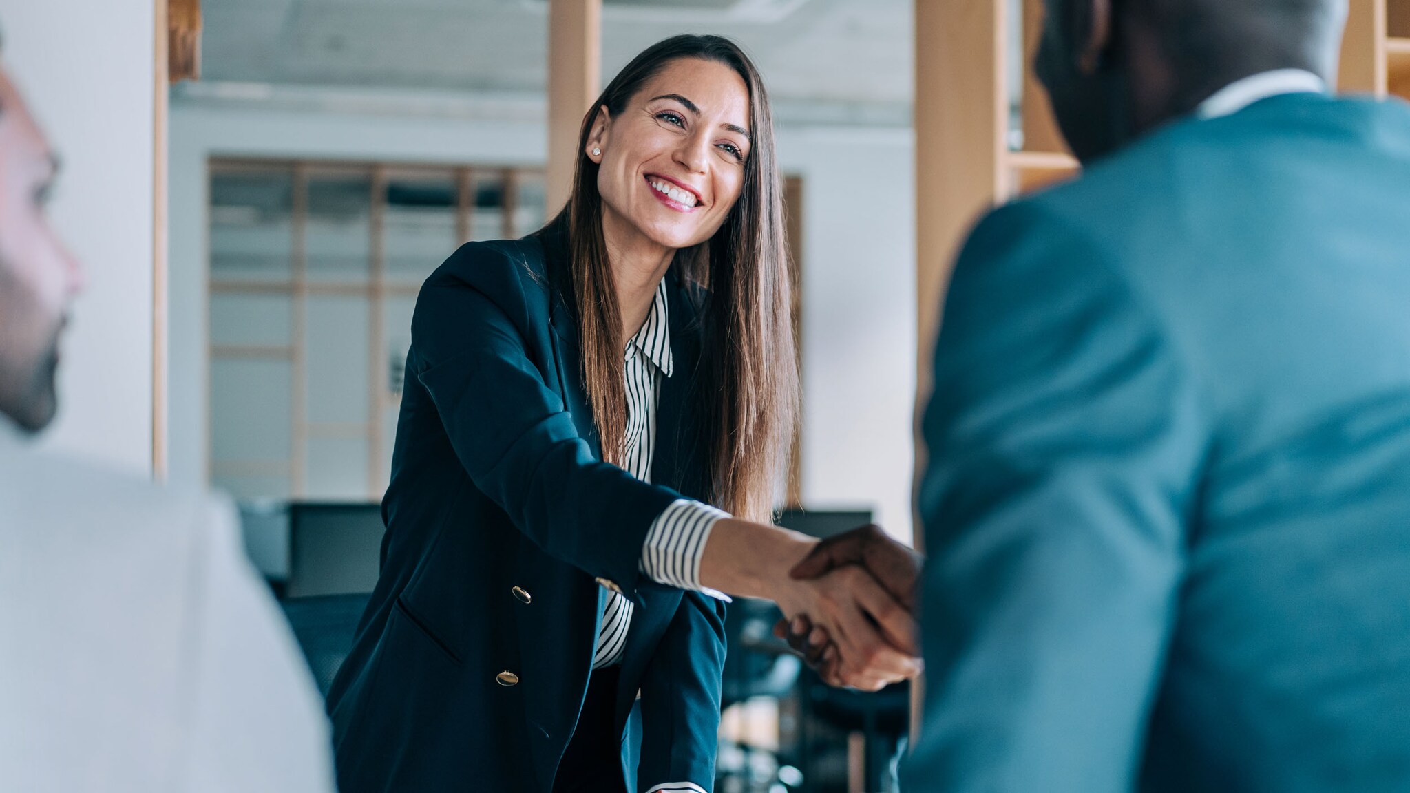 Eine lächelnde Geschäftsfrau im blauen Blazer und ein Mann im Anschnitt aus der Rückansicht geben sich die Hand in einem Büro.