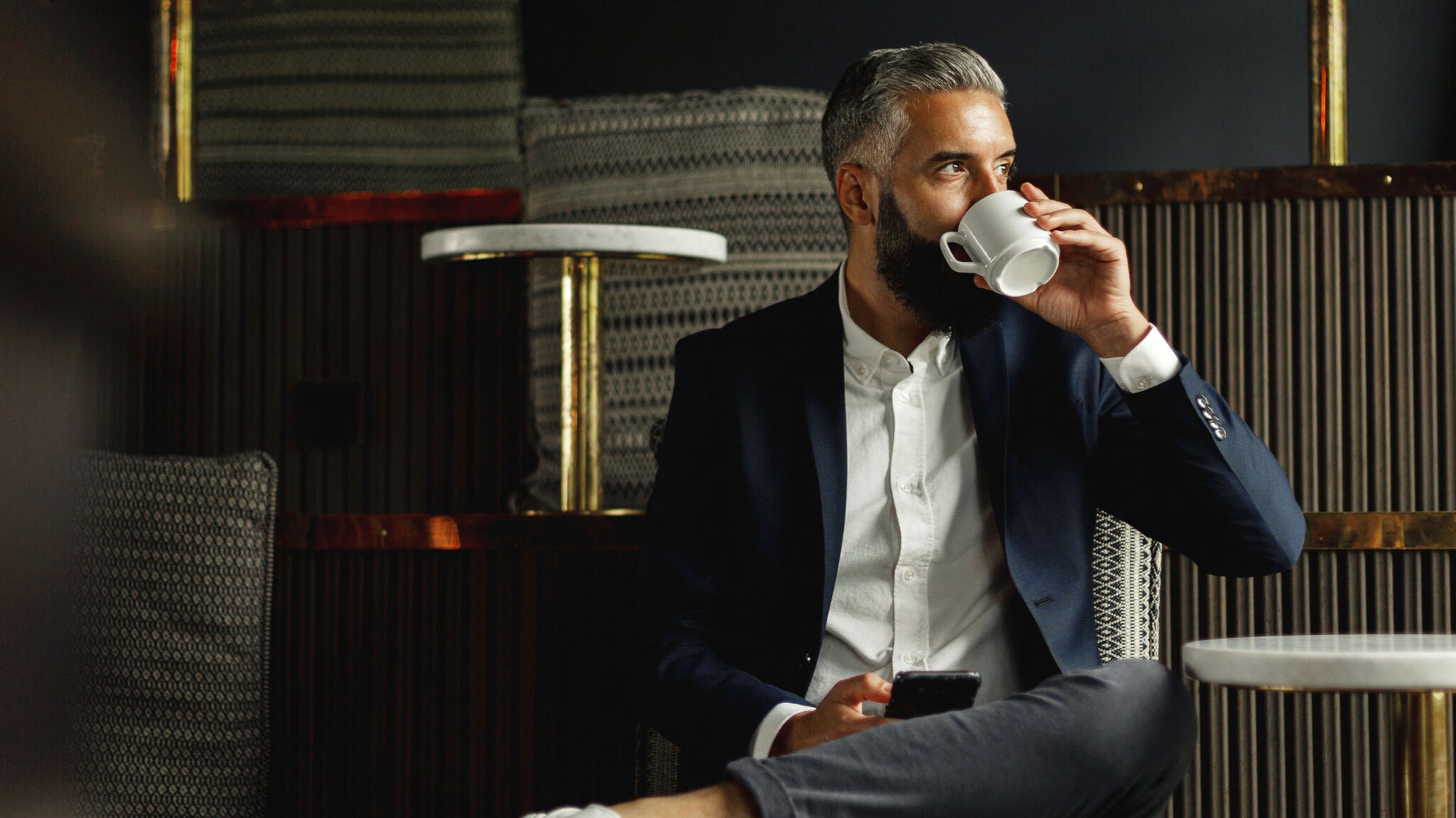 Ein moderner Geschäftsmann sitzt mit Smartphone in der Hand auf einer Sitzempore in einer Lounge und trinkt an einer Kaffeetasse.