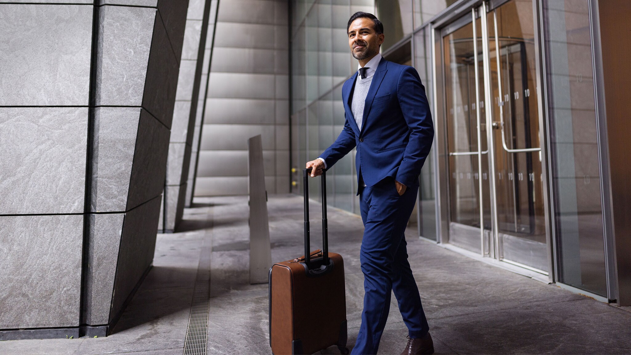 Ein elegant gekleideter Geschäftsmann im blauen Anzug geht mit braunem Trolley vor einem Büroeingang.