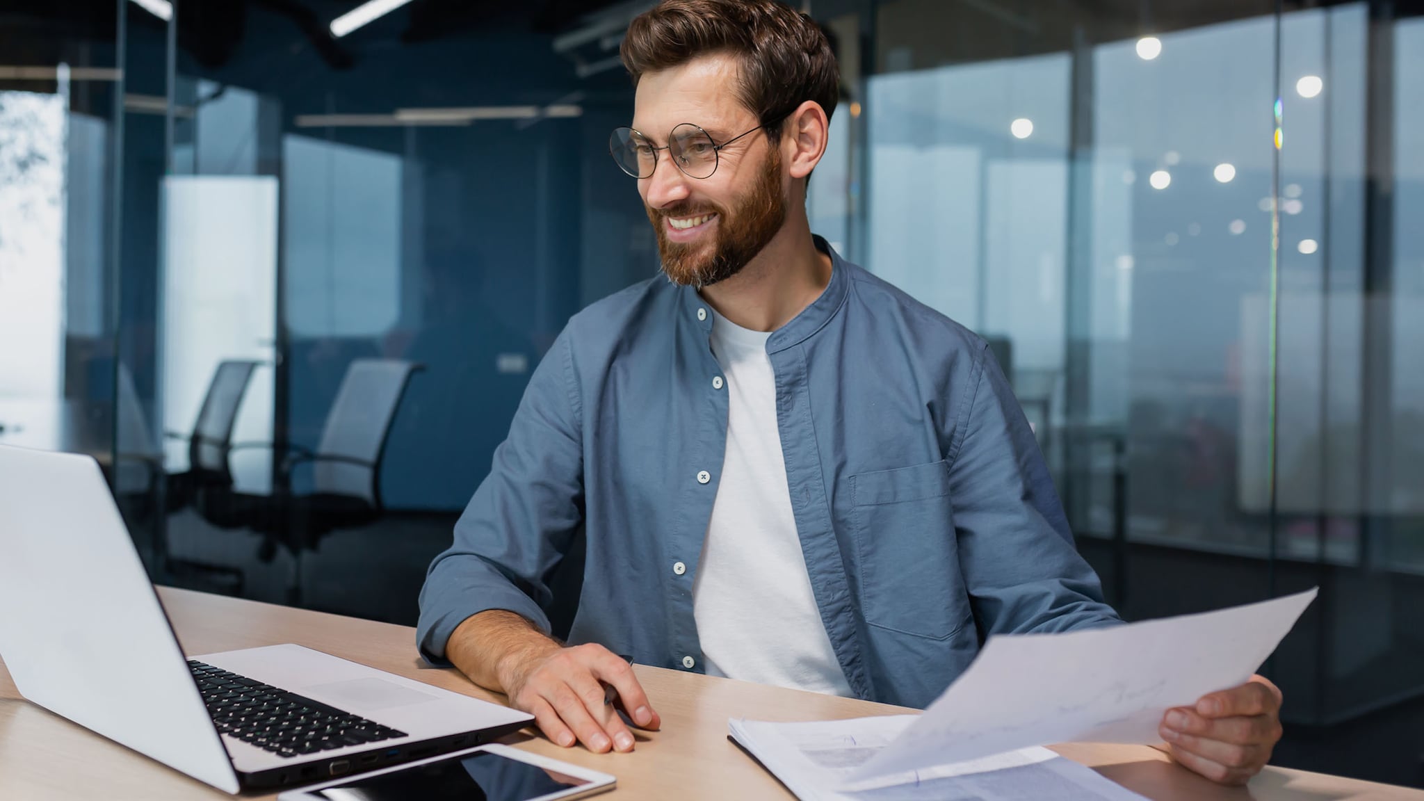 Ein lächelnder Mann sitzt an einem Schreibtisch in einem modernen Büro bei der Arbeit an einem Laptop mit Dokumenten in der Hand.