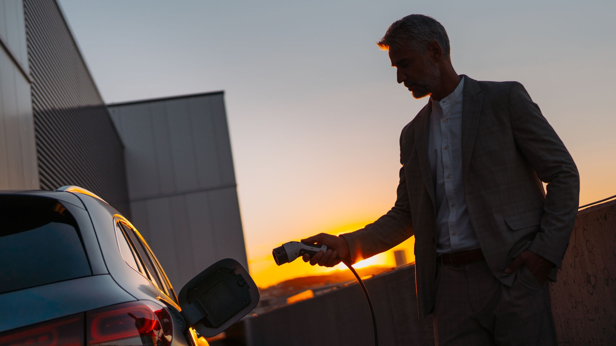 Silhouette eines Mannes mit Ladestecker in der Hand an einem Auto an einem modernen Gebäude bei Sonnenuntergang.