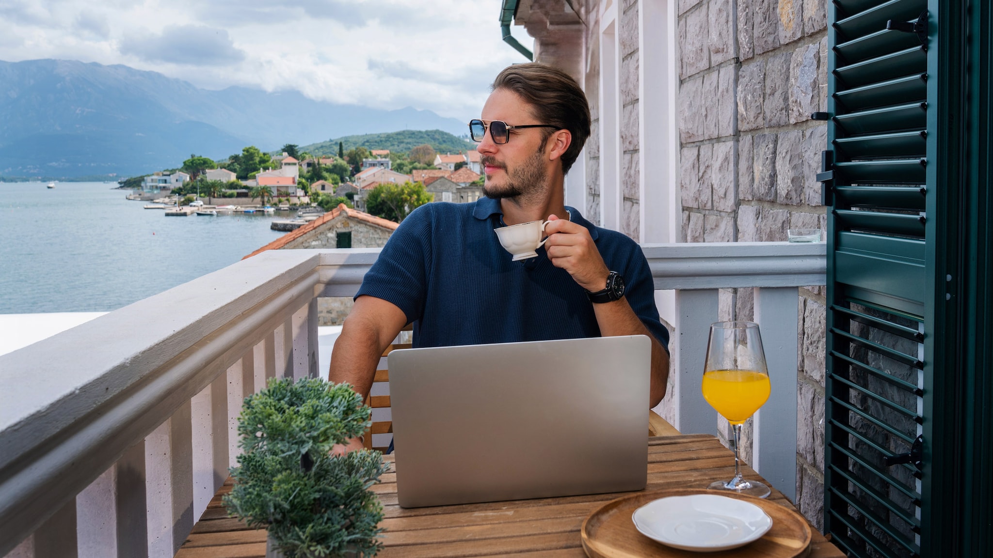 Ein leger gekleideter Mann mit Sonnenbrille und Kaffeetasse in der Hand sitzt hinter einem aufgeklappten Laptop an einem Tisch auf einem Balkon an einer Küste und blickt aufs Meer.