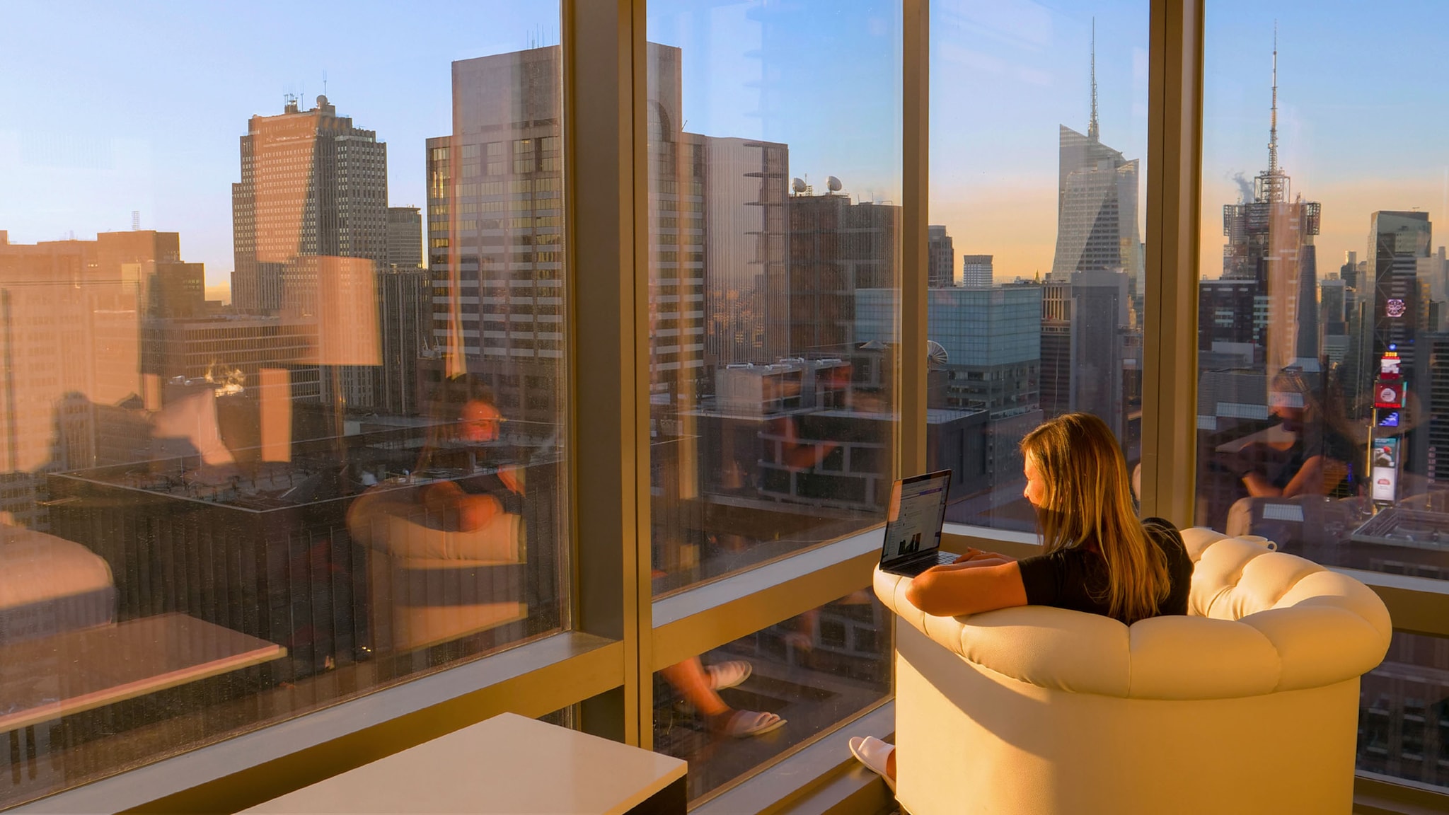 Rückansicht einer Frau, die in einem Sessel an einem Laptop arbeitet, vor einem Panoramafenster mit Blick auf eine Skyline bei Sonnenuntergang.
