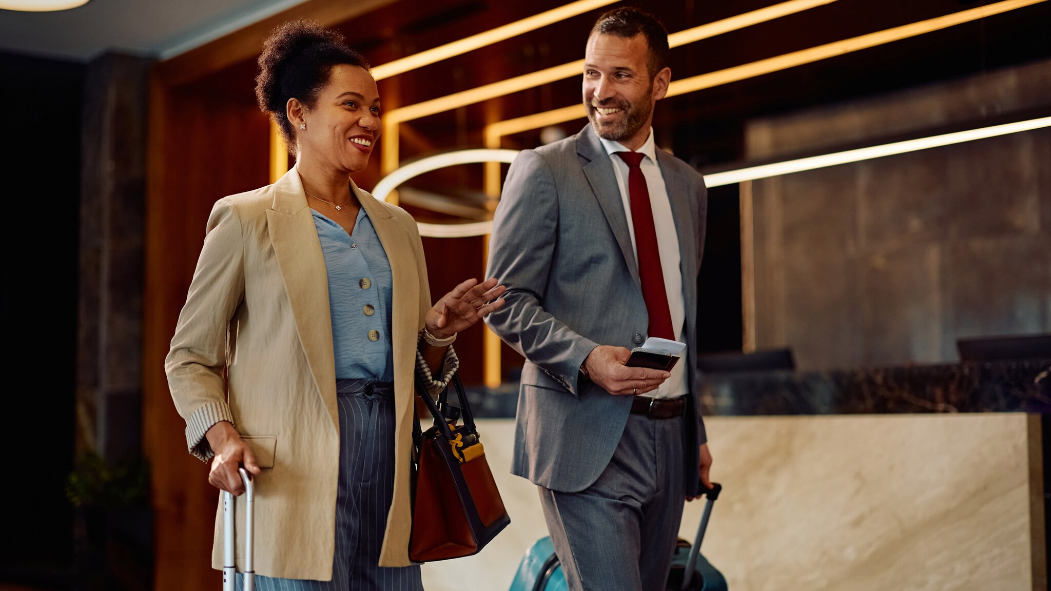 Eine Frau und ein Mann in Businesskleidung gehen mit Rollkoffern an einer Hotelrezeption gut gelaunt nebeneinanderher.