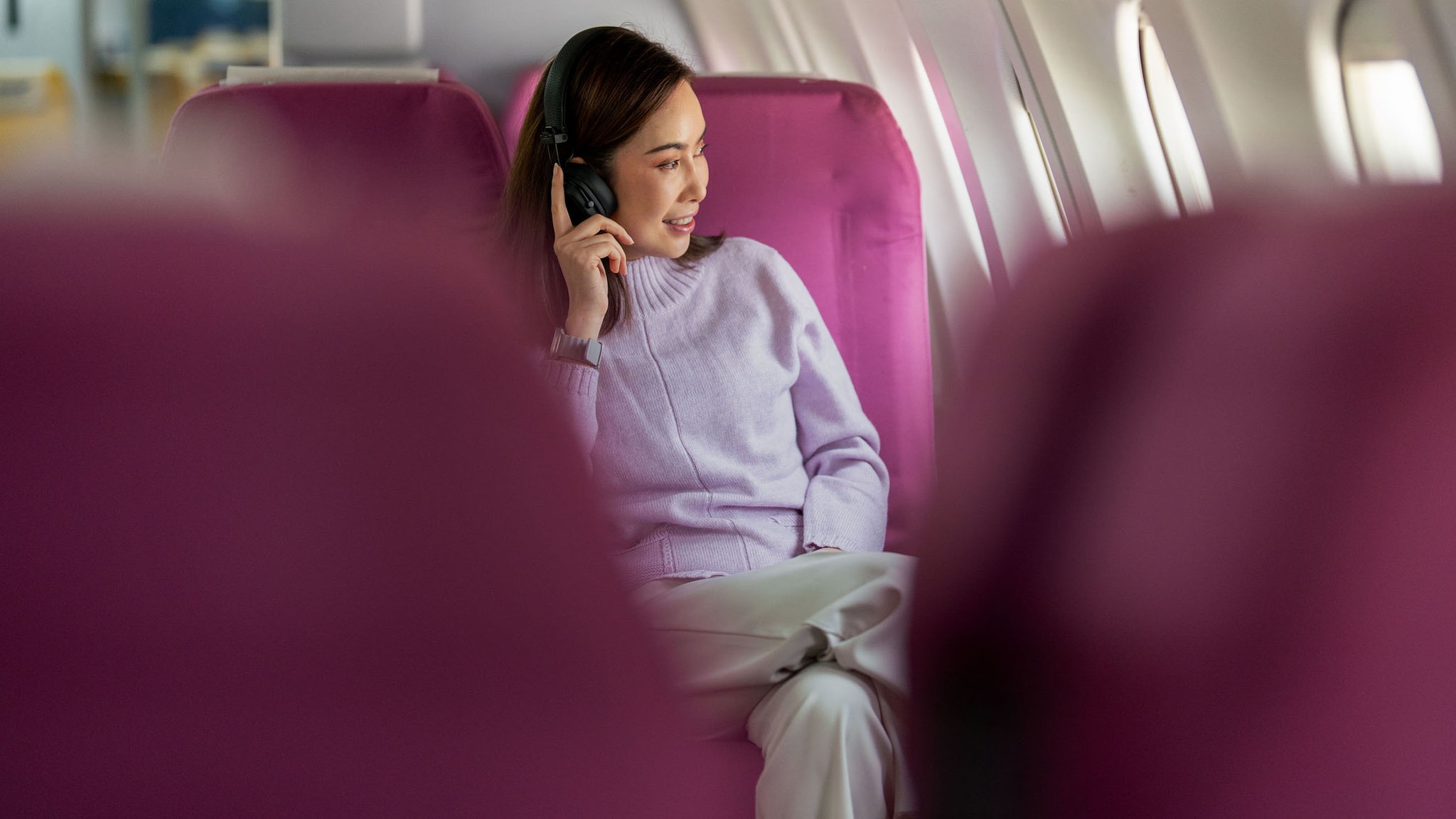 Eine Frau sitzt mit Kopfhörern entspannt auf einem Sitzplatz mit magentafarbenen Bezug in einem Flugzeug und schaut aus dem Fenster, im Vordergrund leere Sitzplätze in Unschärfe.