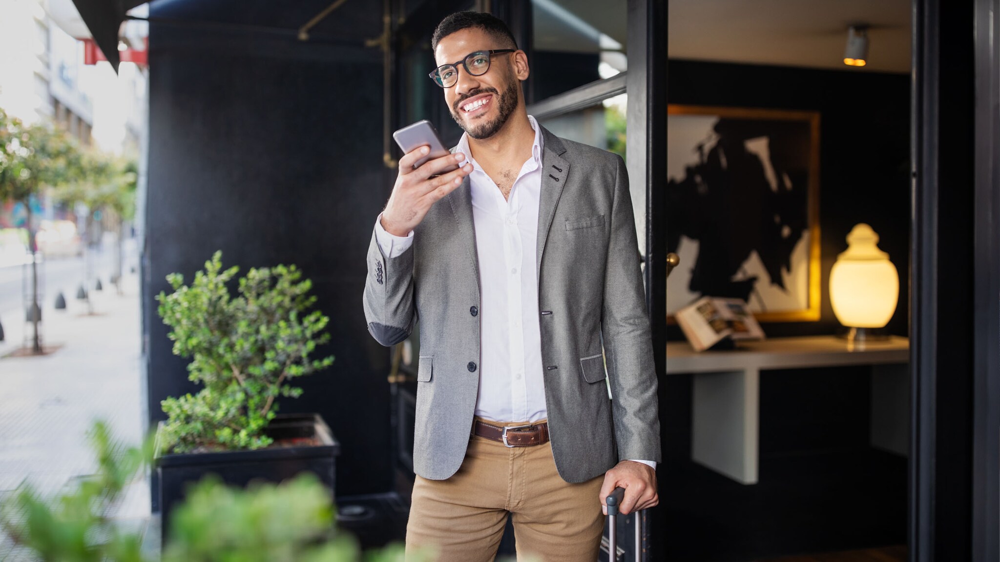 Ein moderner Geschäftsmann steht mit Trolley im Eingangsbereich eines Hotels und spricht gut gelaunt in ein Smartphone in seiner Hand.