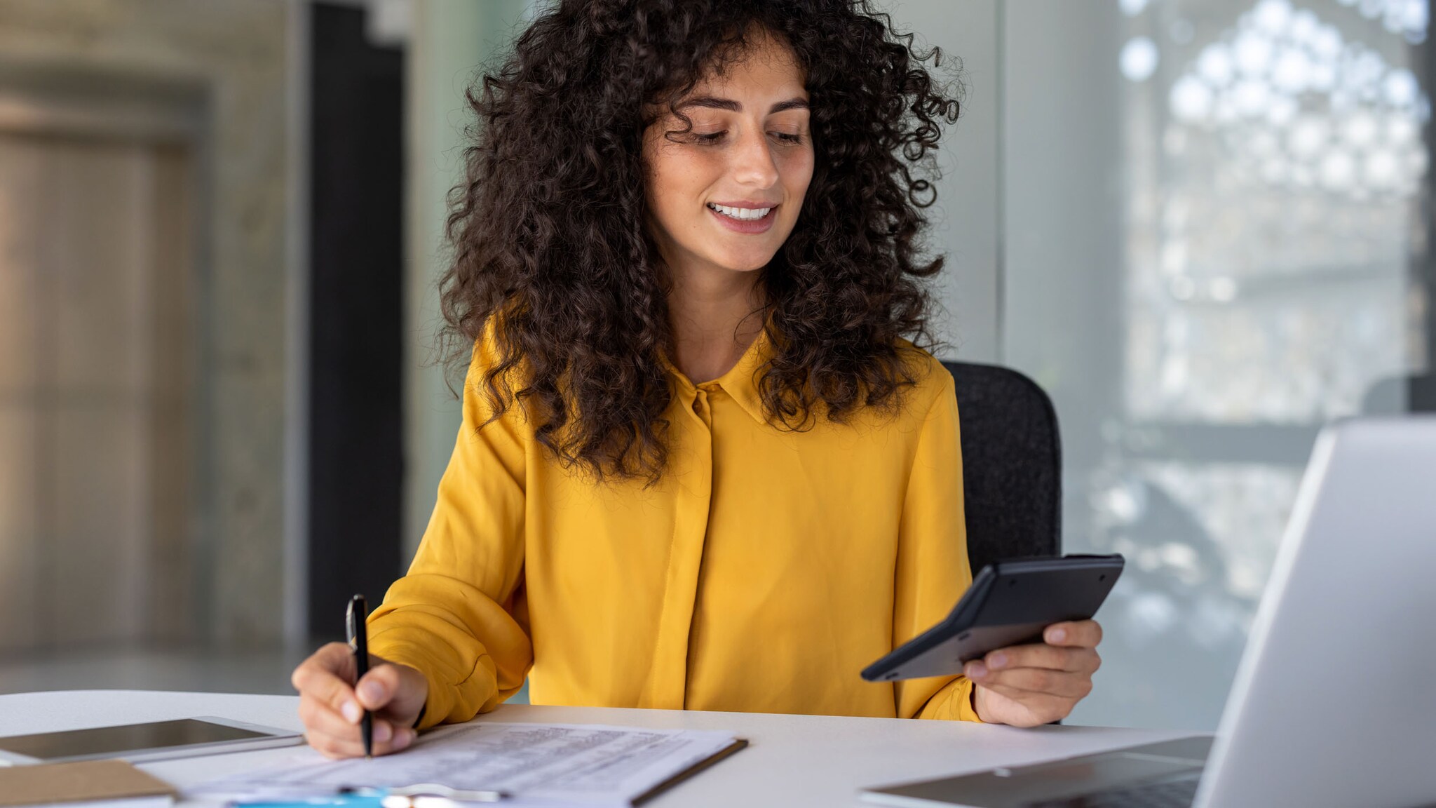Eine Frau in gelber Bluse bearbeitet Dokumente an einem Schreibtisch, während sie einen Taschenrechner in der Hand hält.