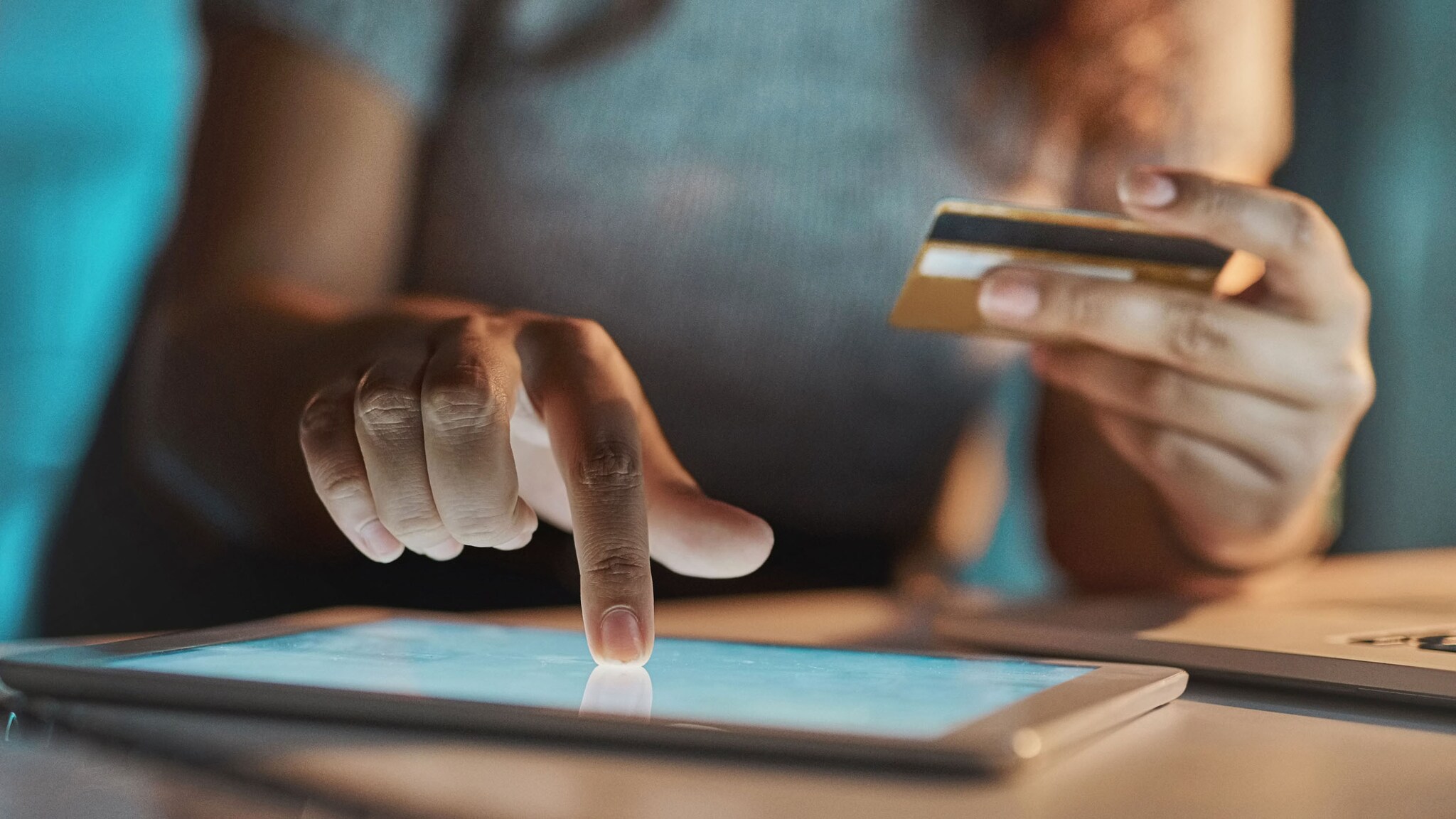 Nahaufnahme von zwei Händen einer Person in einer abgedunkelter Raumumgebung, die eine Kreditkarte hält, während sie ein beleuchtetes Tablet auf einem Tisch bedient.