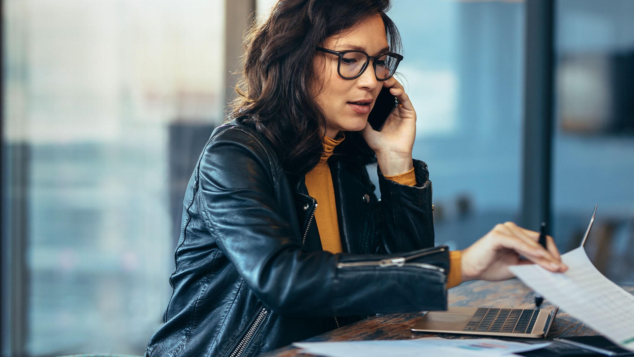 Eine Frau in Lederjacke sitzt an einem Schreibtisch vor einem Laptop und greift nach Dokumenten, während sie mit einem Smartphone telefoniert in einem Büro.