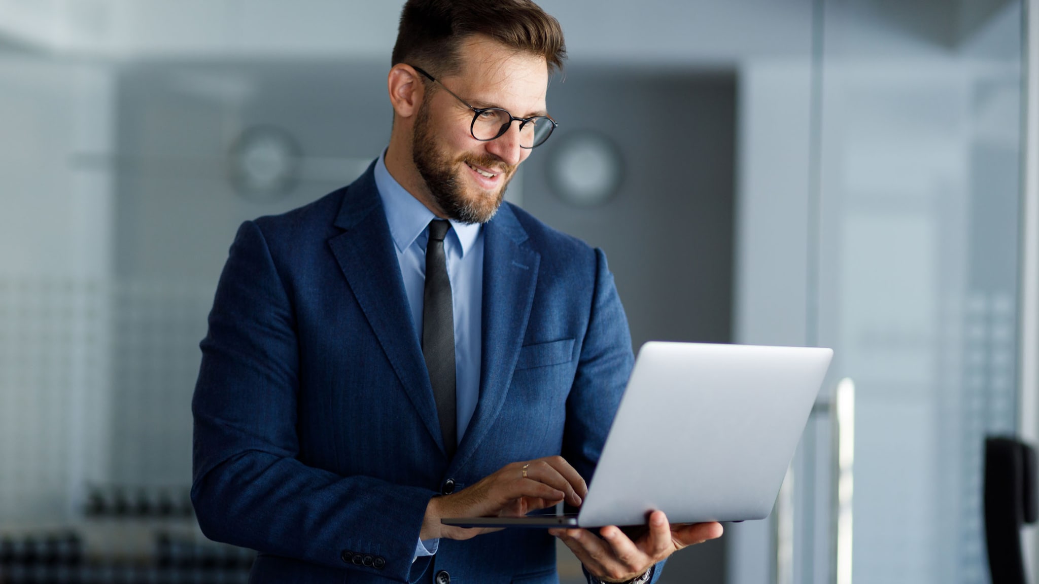 Ein Mann im blauen Anzug in einem Büro arbeitet stehend an einem Laptop in der Hand.