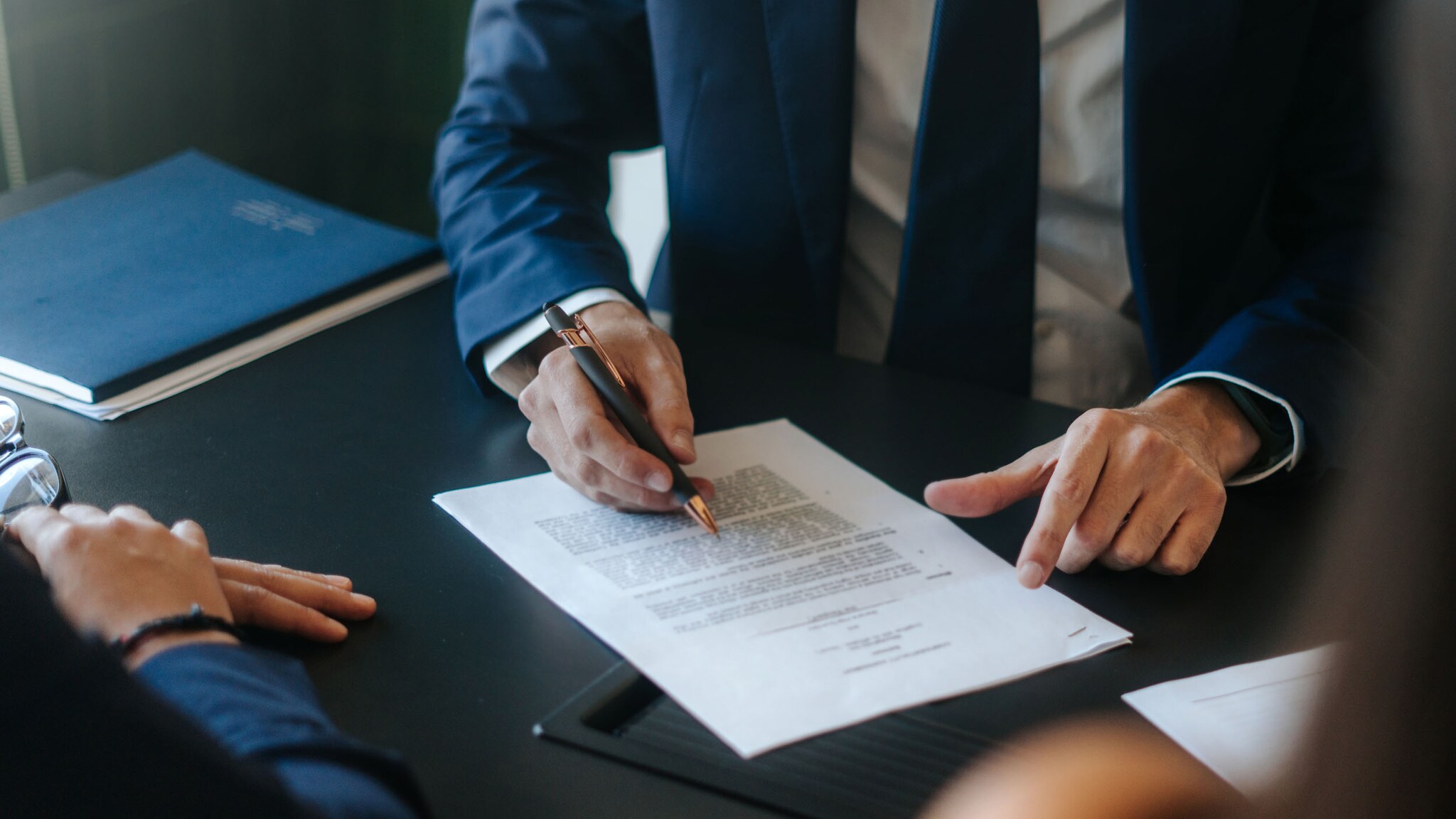 Nahaufnahme eines Dokumentes auf einem Tisch, das von einem Mann im blauen Anzug mit Krawatte unterschrieben wird, im Vordergrund zwei Personen im Anschnitt.
