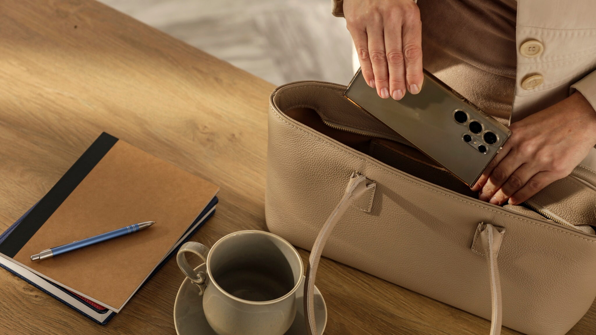 Eine Person in einem hellen Blazer steckt ein Smartphone in eine beige Lederhandtasche auf einem Holztisch.