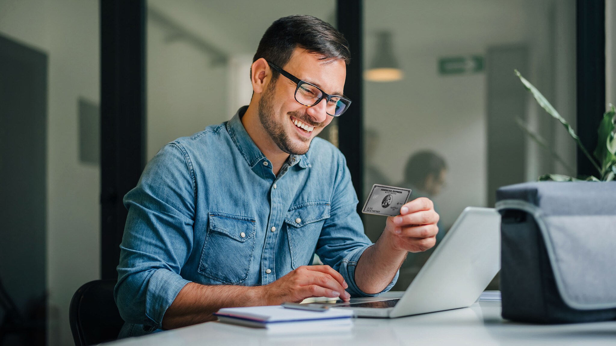 Ein lächelnder Mann im Jeanshemd bedient ein Laptop an einem Tisch in einem Büro und hält eine silberne Business Kreditkarte von American Express in der Hand.