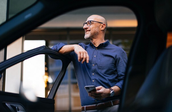 Ein Mann im blauen Hemd stützt einen Arm auf eine geöffnete Autotür, hält ein Smartphone in der Hand und blickt lächelnd zur Seite.