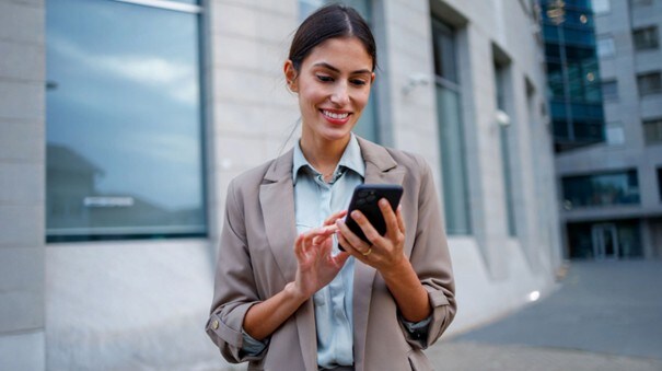 Eine Frau in Business-Kleidung bedient lächelnd ein Smartphone in ihrer Hand vor einem Bürogebäude.