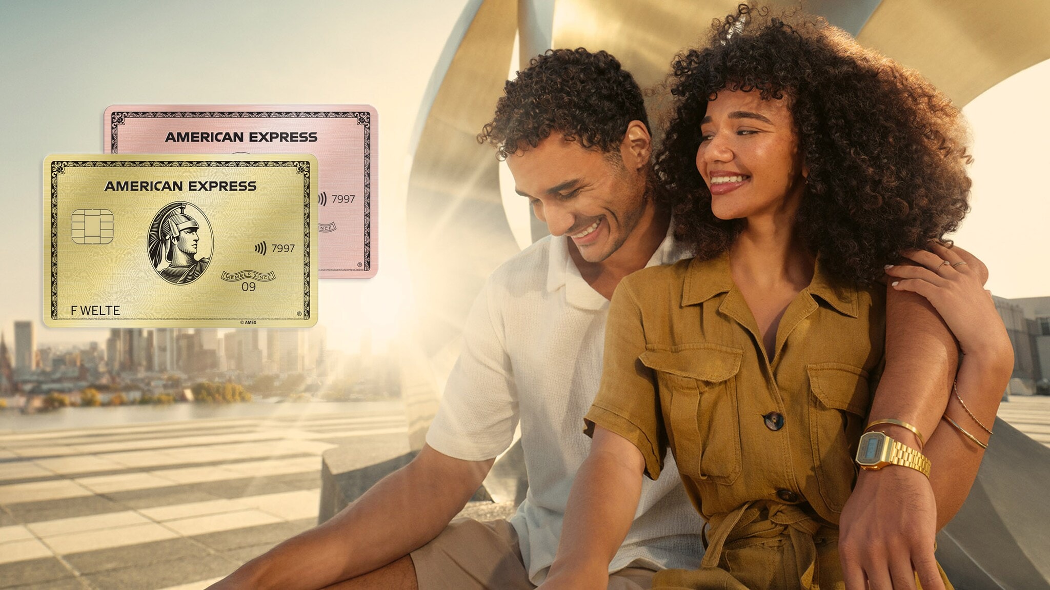 Ein Mann und eine Frau sitzen in einer urbanen Umgebung und lächeln sich an. Im Vordergrund sieht man zwei Kreditkarten von American Express.