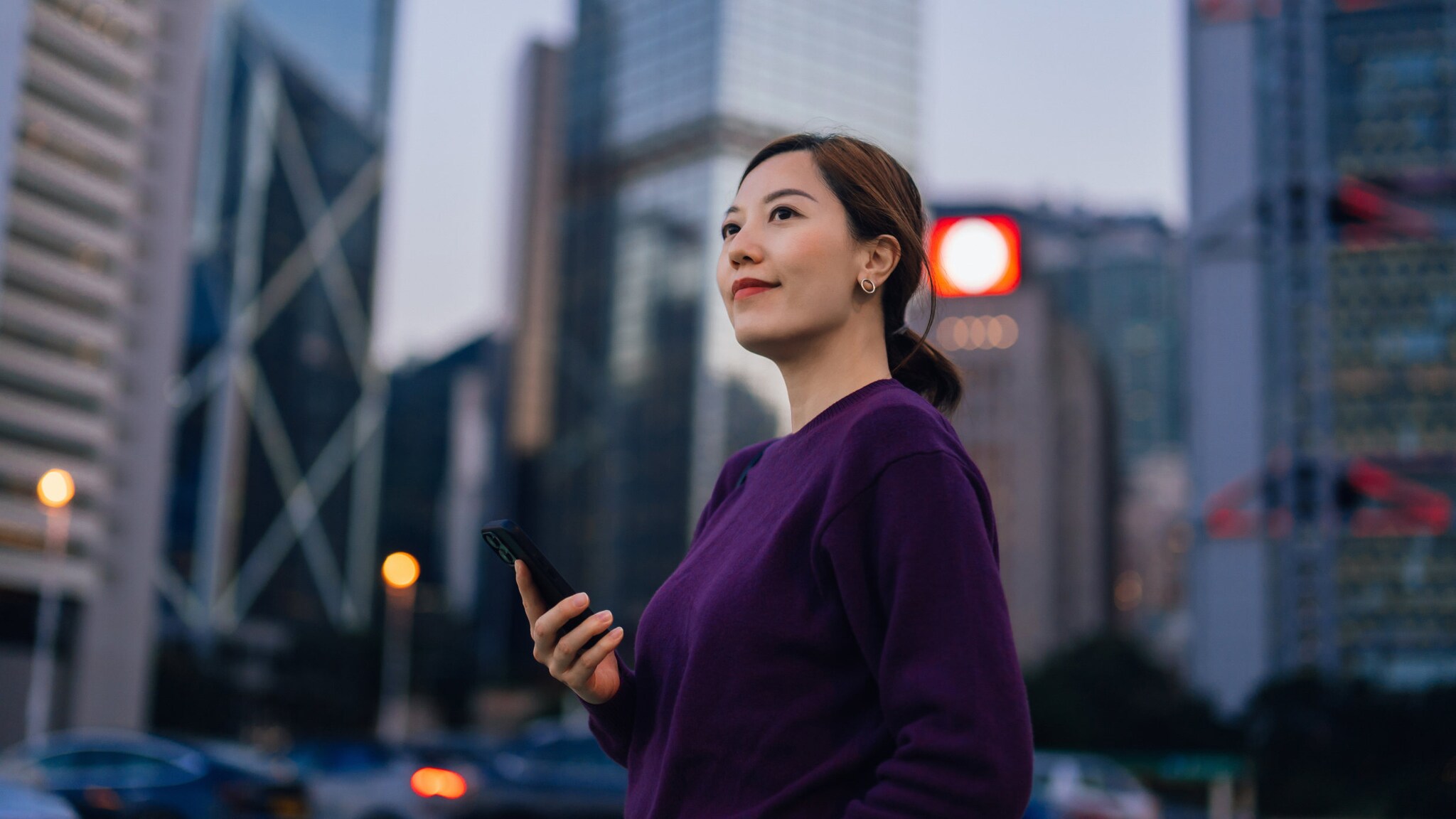 Selbstbewusste junge Geschäftsfrau mit Smartphone in der Hand, die in der Innenstadt zwischen Hochhäusern steht und in die Ferne blickt. 