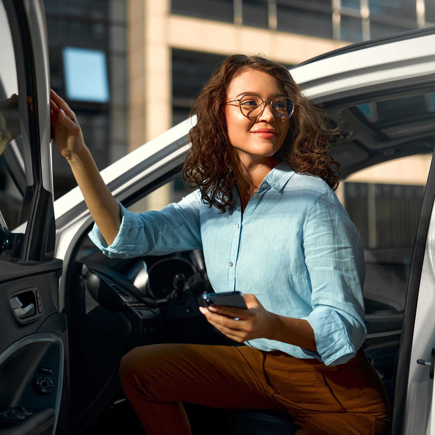 Eine junge Frau in hellblauer Bluse steigt mit Smartphone in der Hand aus einem Auto aus.