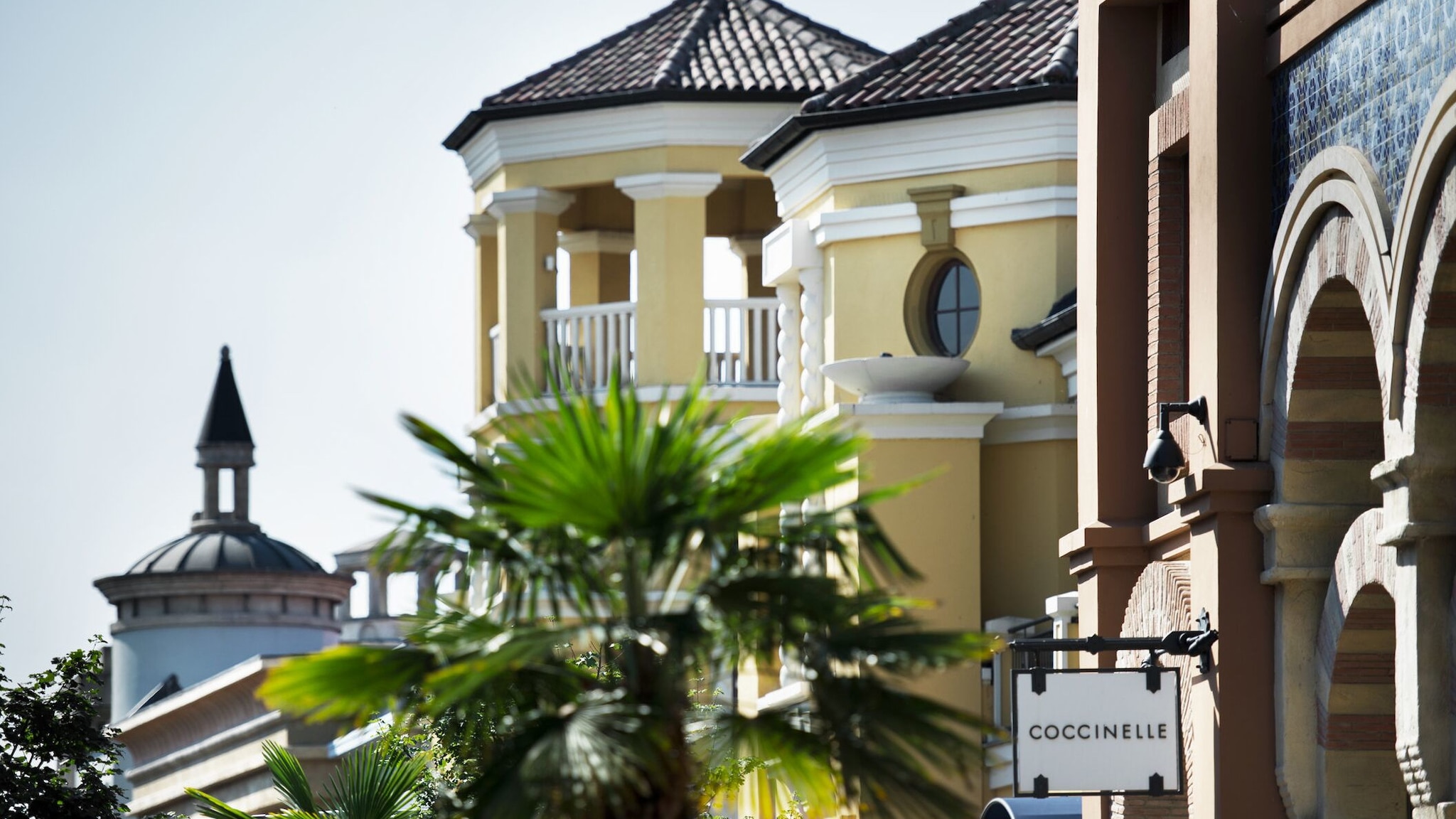 Close-up von turm- und villenähnlichen Gebäuden der The Bicester Collection mit grüner Palme und Boutique-Schild im Vordergrund. Close-up von turm- und villenähnlichen Gebäuden der The Bicester Collection mit grüner Palme und Boutique-Schild im Vordergrund.