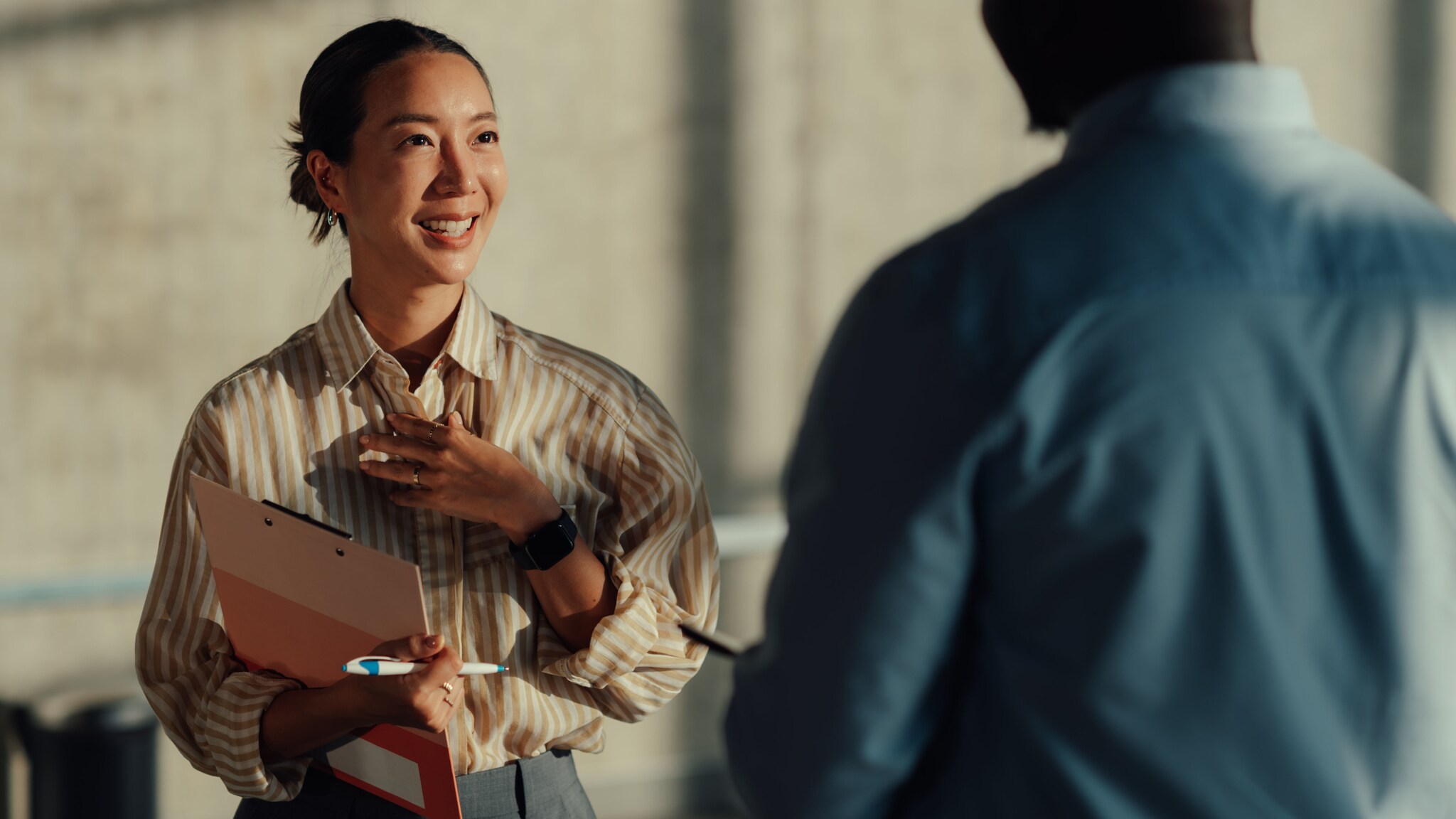 amex-sbs-interkulturelle-kommunikation-frau-klemmbrett Eine junge lächelnde Frau in gestreifter Bluse und Klemmbrett im Arm kommuniziert mit einem Mann im blauen Hemd in der Rückansicht.