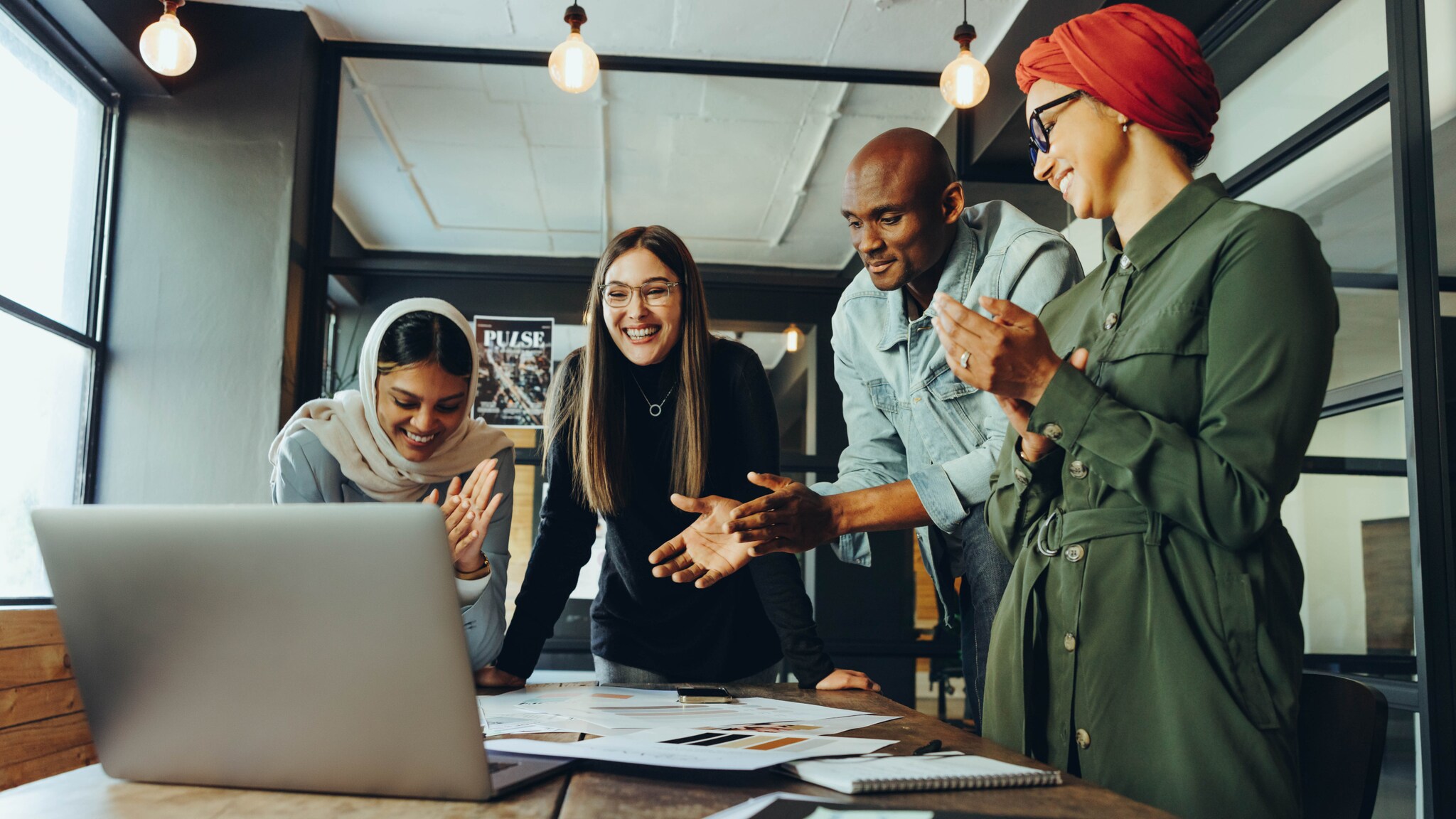 Vier Personen multiethnischer Herkunft interagieren lächelnd mit einem aufgeklappten Laptop auf einem Schreibtisch in einem Büro.