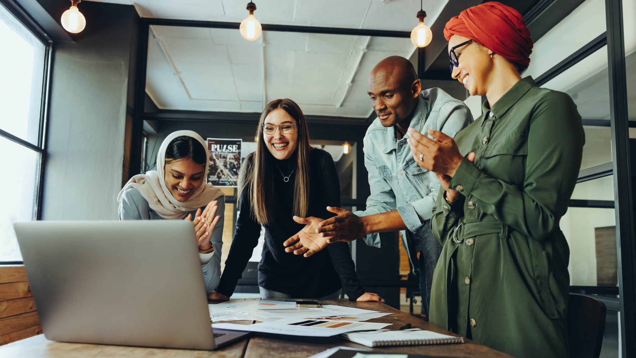amex-sbs-interkulturelle-kommunikation-laptop-buero Vier Personen multiethnischer Herkunft interagieren lächelnd mit einem aufgeklappten Laptop auf einem Schreibtisch in einem Büro.