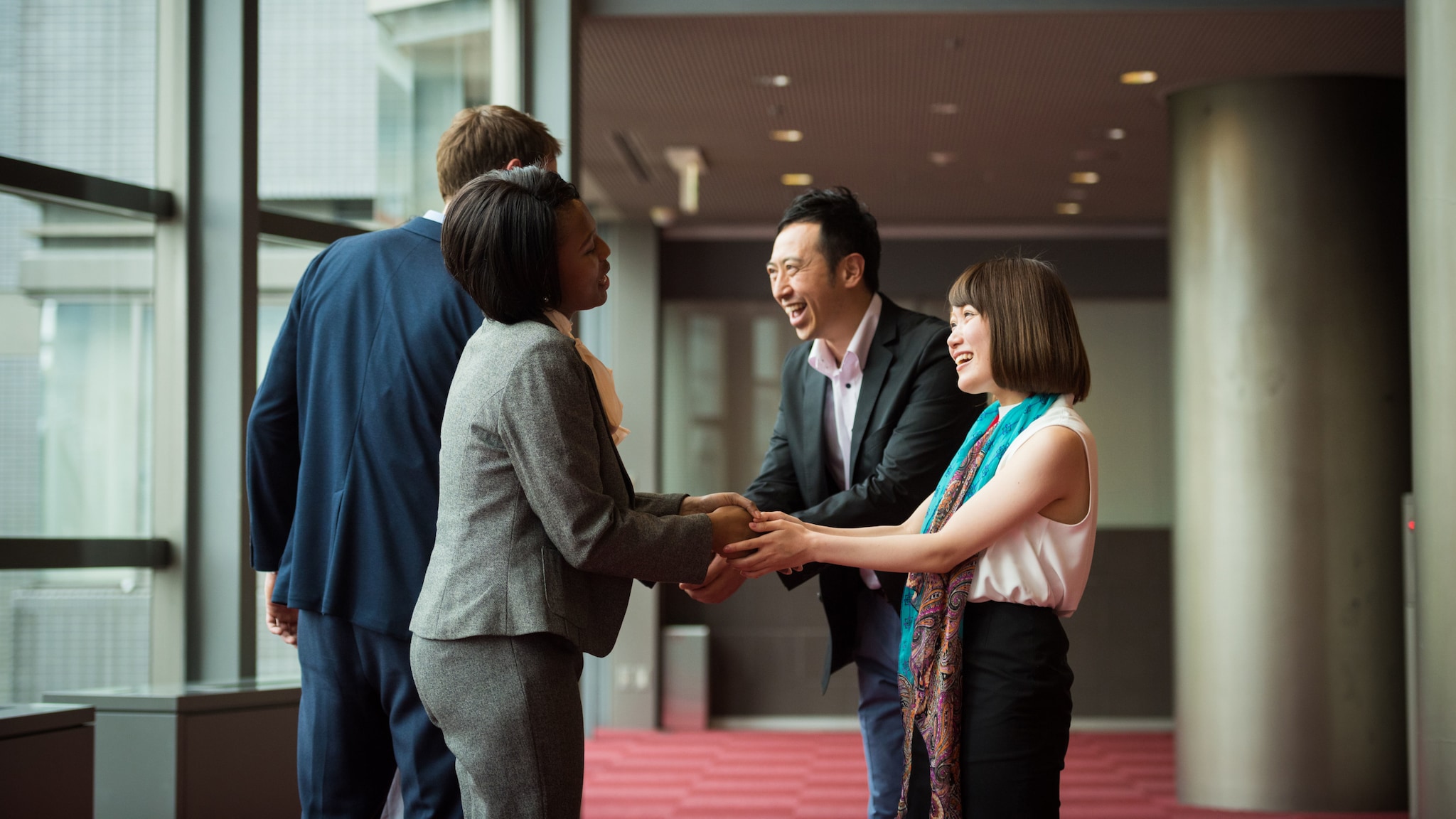 amex-sbs-interkulturelle-kommunikation-lobby-vier-personen Zwei Männer und zwei Frauen multiethnischer Herkunft in Business-Kleidung geben sich lächelnd die Hände in einer Lobby.