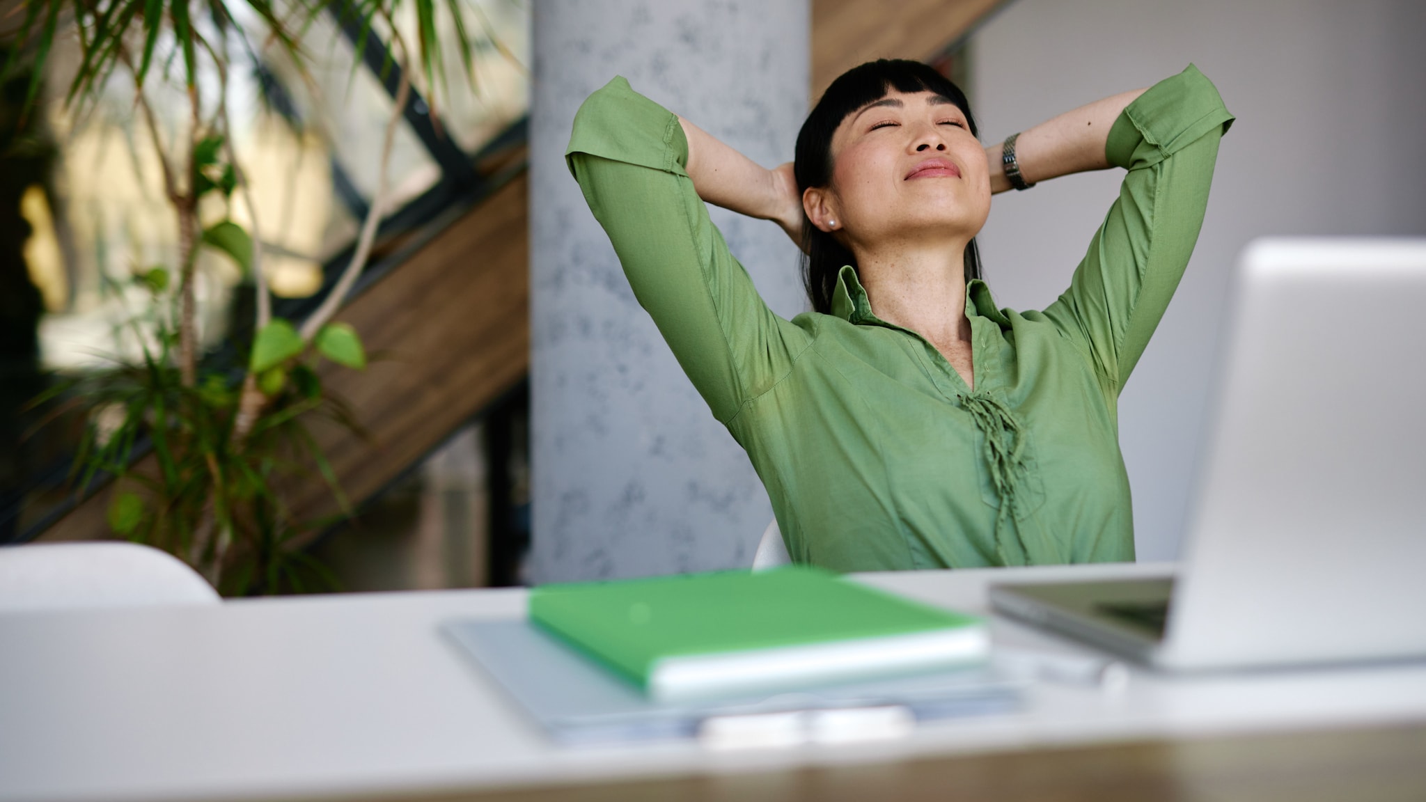 amex-sbs-mentale-staerke-frau-gruener-bluse Eine Frau in grüner Bluse lehnt mit geschlossenen Augen und verschränkten Armen hinter dem Kopf entspannt zurück auf einem Stuhl an einem Schreibtisch in einem Büro.
