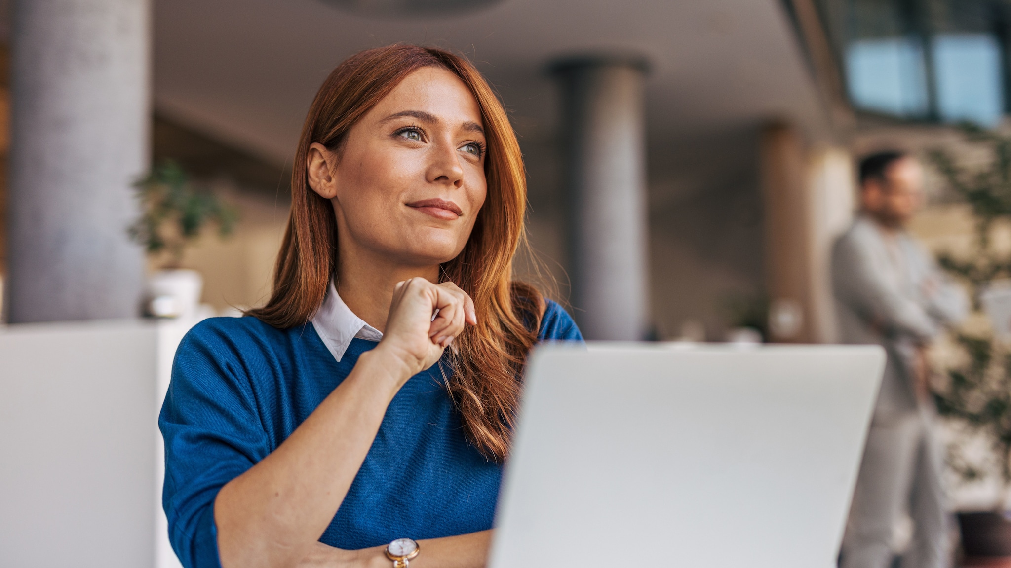 amex-sbs-mentale-staerke-frau-laptop Nahaufnahme einer nachdenklichen, selbstbewusst schauenden Frau im blauen Pullover hinter einem aufgeklappten Laptop in einem Großraumbüro.