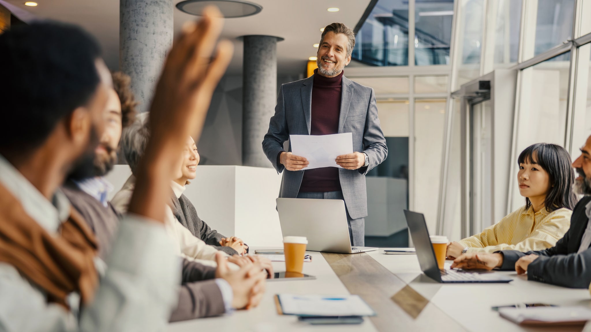 amex-sbs-mentale-staerke-konferenztisch-meeting Ein lächelnder Mann im grauen Sakko steht mit Arbeitsunterlagen in den Händen an einem Konferenztisch während eines Meetings und blickt zu einem Mitarbeitenden, der die Hand hebt in einem Großraumbüro.