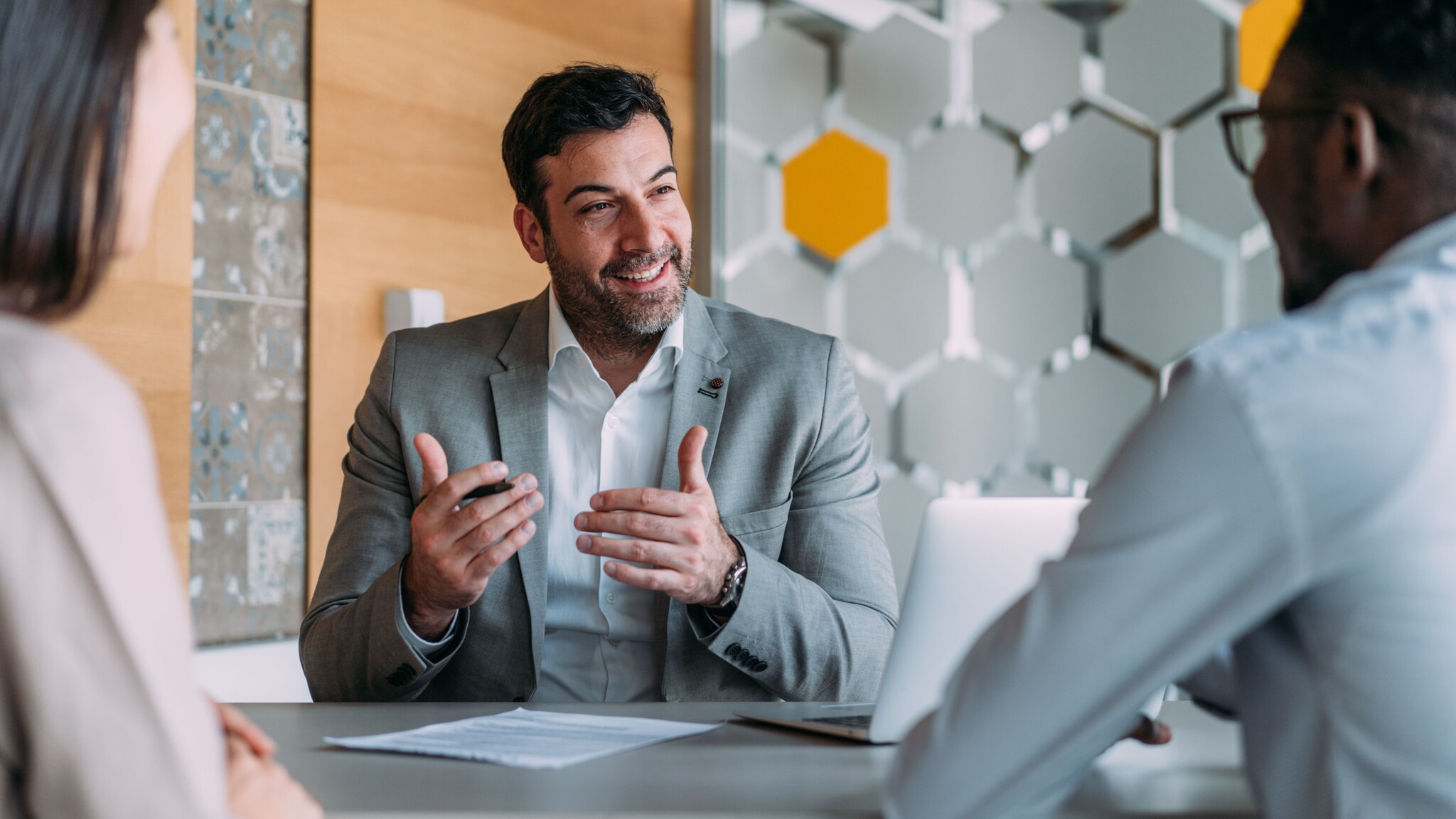 amex-sbs-gespraechsfuehrung-geschaeftsmann-schreibtisch Ein Geschäftsmann im grauen Sakko gestikuliert mit den Händen während eines Meetings mit zwei weiteren Personen in der Rückansicht an einem Schreibtisch in einem Büro.