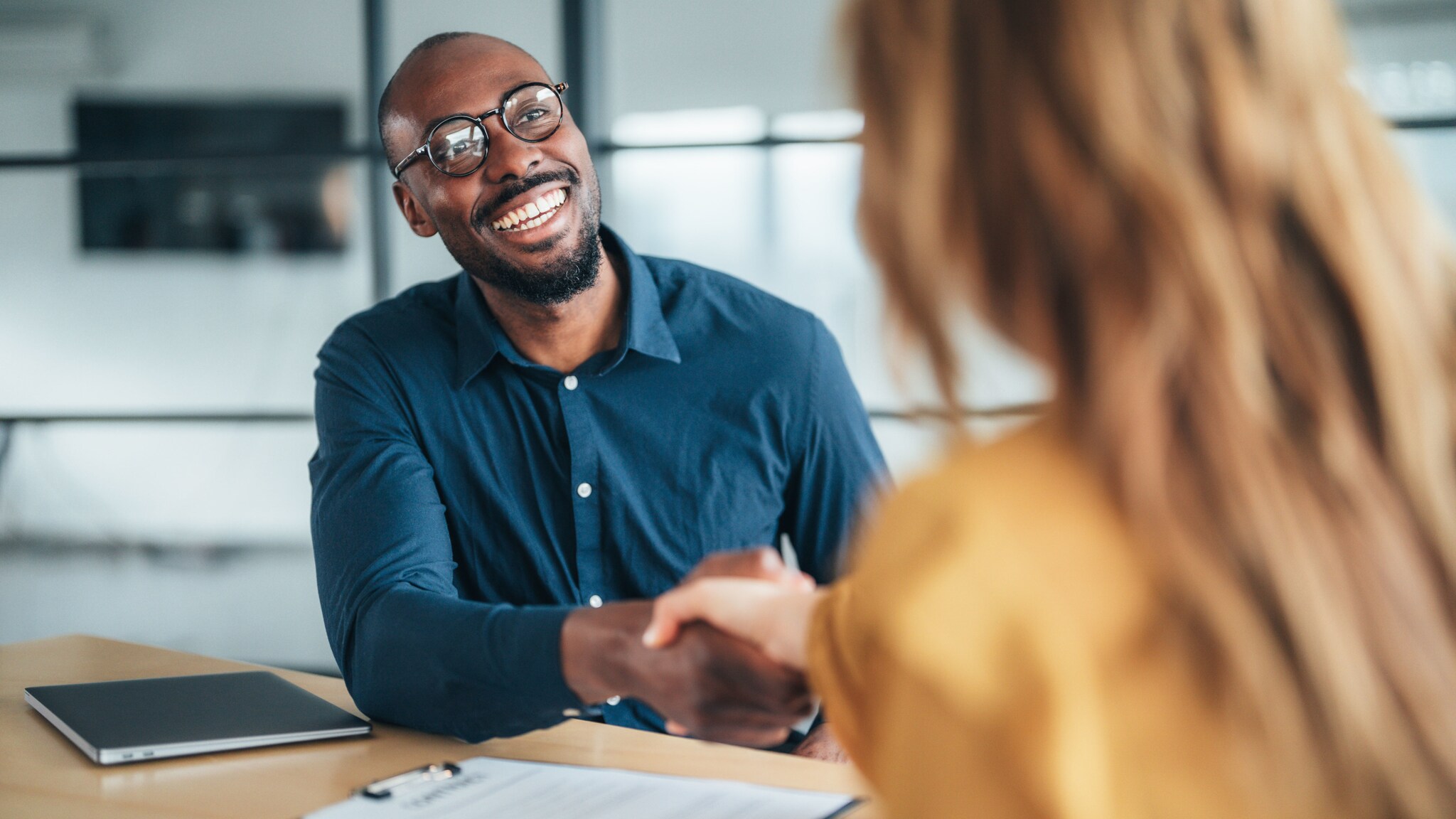 Ein offen lachender Mann im blauen Hemd und eine Frau in der Rückansicht geben sich die Hände an einem Schreibtisch mit Geschäftsunterlagen in einem Büro.