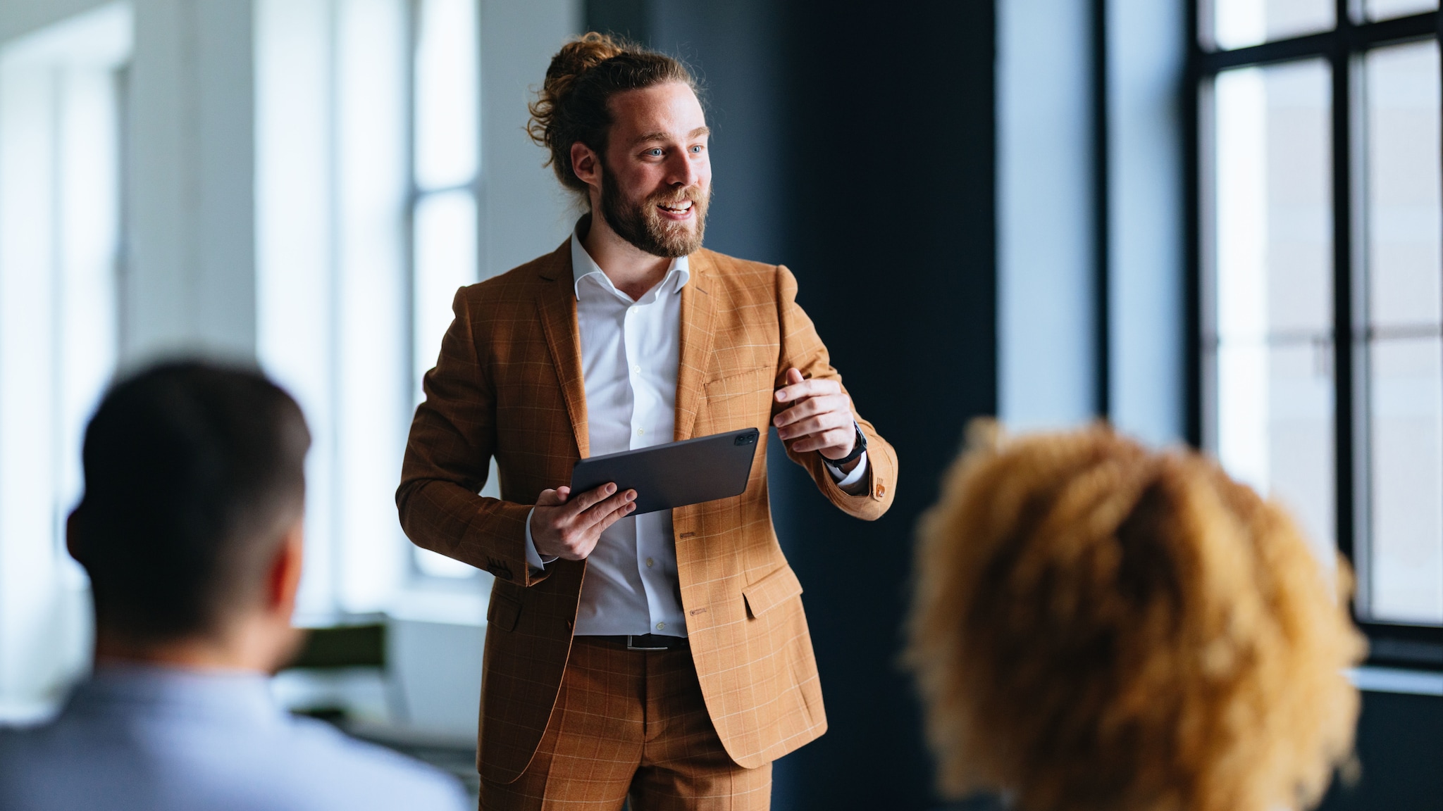 amex-sbs-business-dresscode-herren-mann-dutt-frisur Ein Mann mit Dutt-Frisur in einem legeren, ockerfarbenen Anzug referiert mit Tablet in der Hand vor sitzenden Personen in einem Raum.