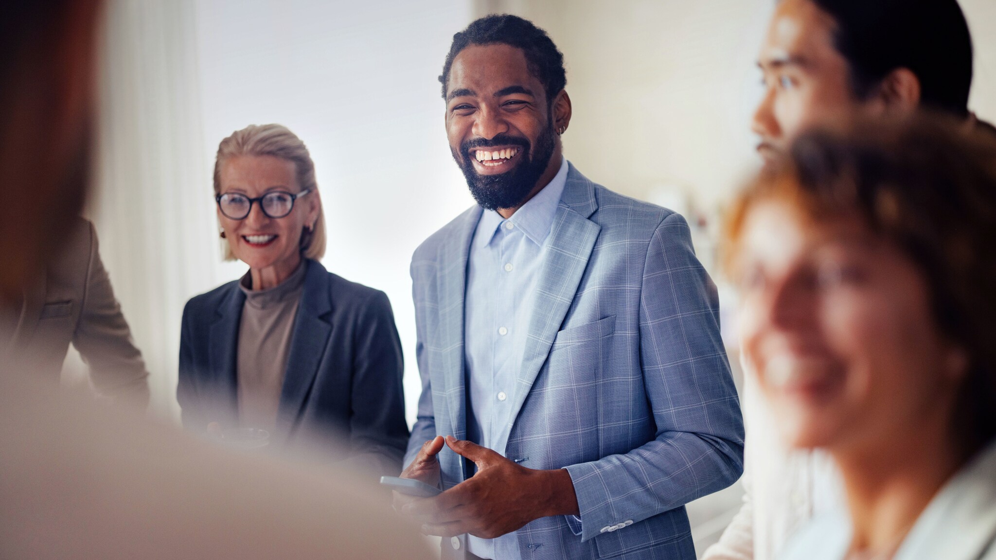 amex-sbs-business-dresscode-herren-mann-karierten-jackett Mehrere Personen stehen beieinander in einem Raum, im Fokus ein lachender Mann im hellblauen Hemd unter einem hellblauen, karierten Jackett.
