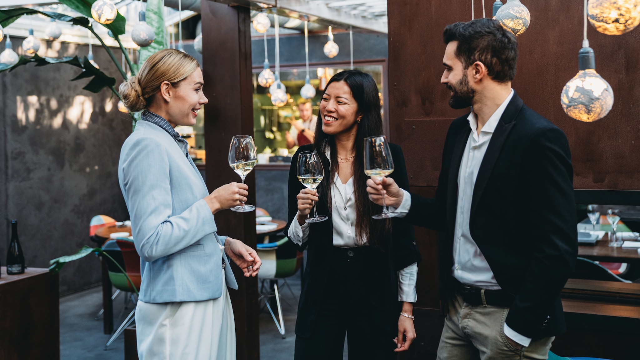amex-sbs-aufbau-von-netzwerken-drei-personen-weinglaesern Drei Personen in eleganten Business-Outfits stehen mit Weingläsern in den Händen beieinander in einem modernen Restaurant.