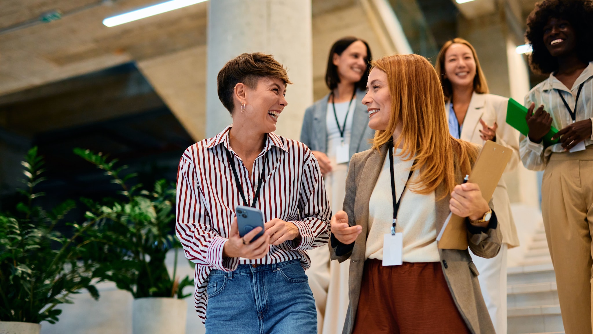 amex-sbs-aufbau-von-netzwerken-fuenf-frauen Fünf Frauen in Business-Outfits mit Arbeitsunterlagen in den Händen und Lanyards um den Hals gehen eine Treppe hinunter in einem Foyer.