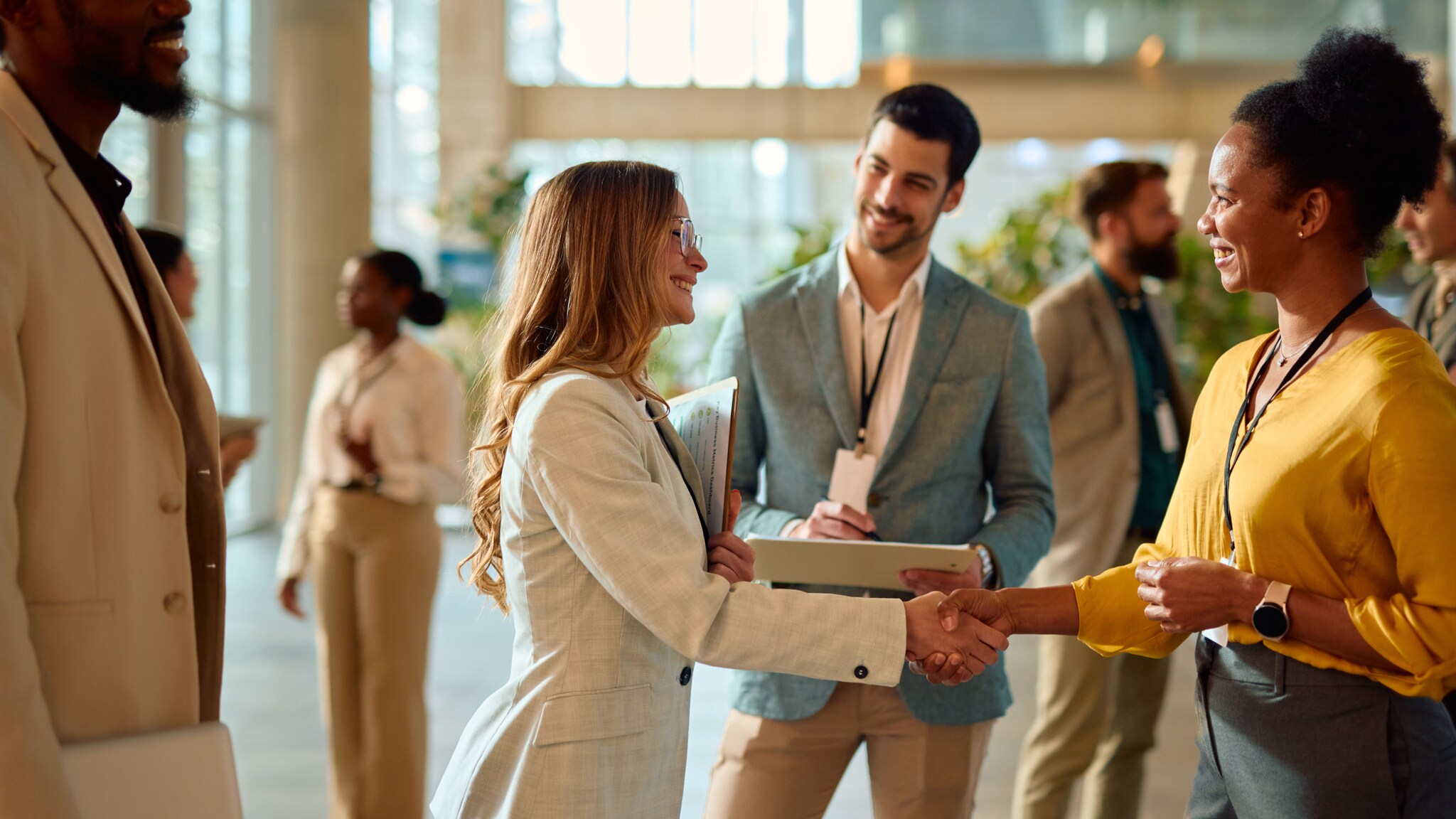 Mehrere Personen in Business-Outfits mit Lanyards um den Hals stehen in einem Foyer, im Vordergrund geben sich zwei Frauen lächelnd die Hand.