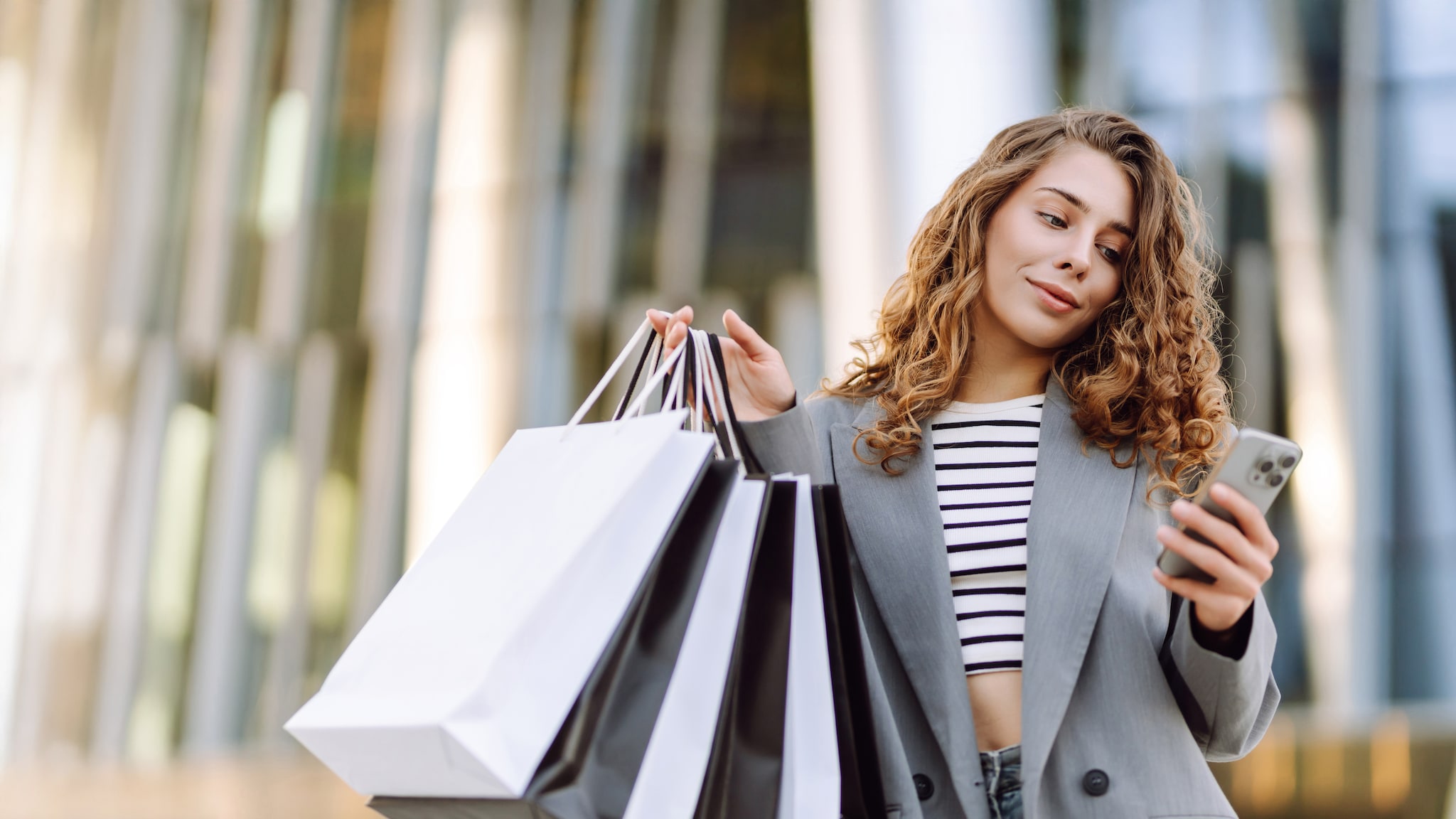 Eine Frau hält Einkaufstüten in der Hand und schaut auf ihr Smartphone.