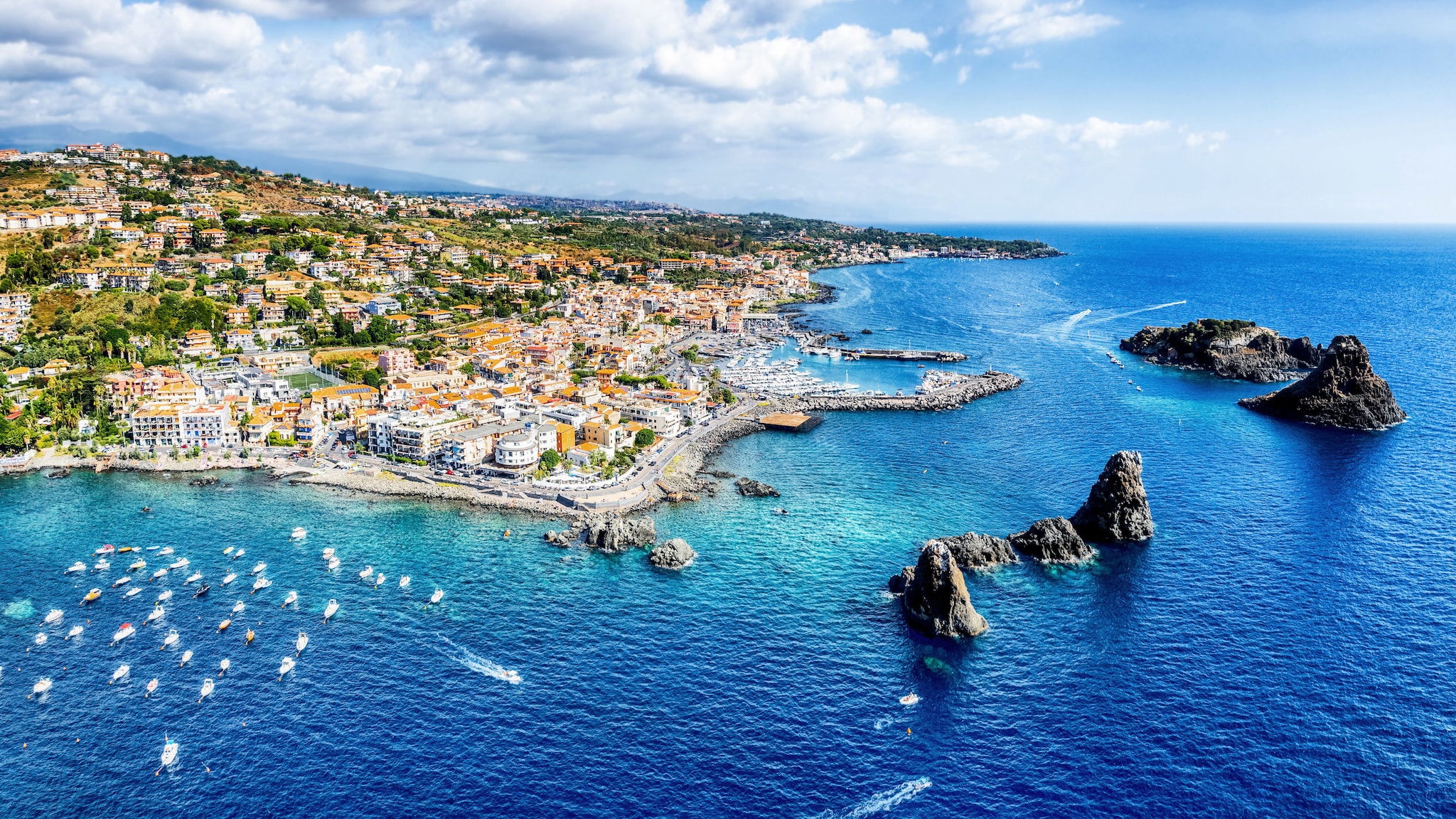 amex-magazin-catania-strand-stadt-kueste Luftaufnahme von einer Stadt am Meer, im Wasser vor der Küste ragen große Felsen empor.