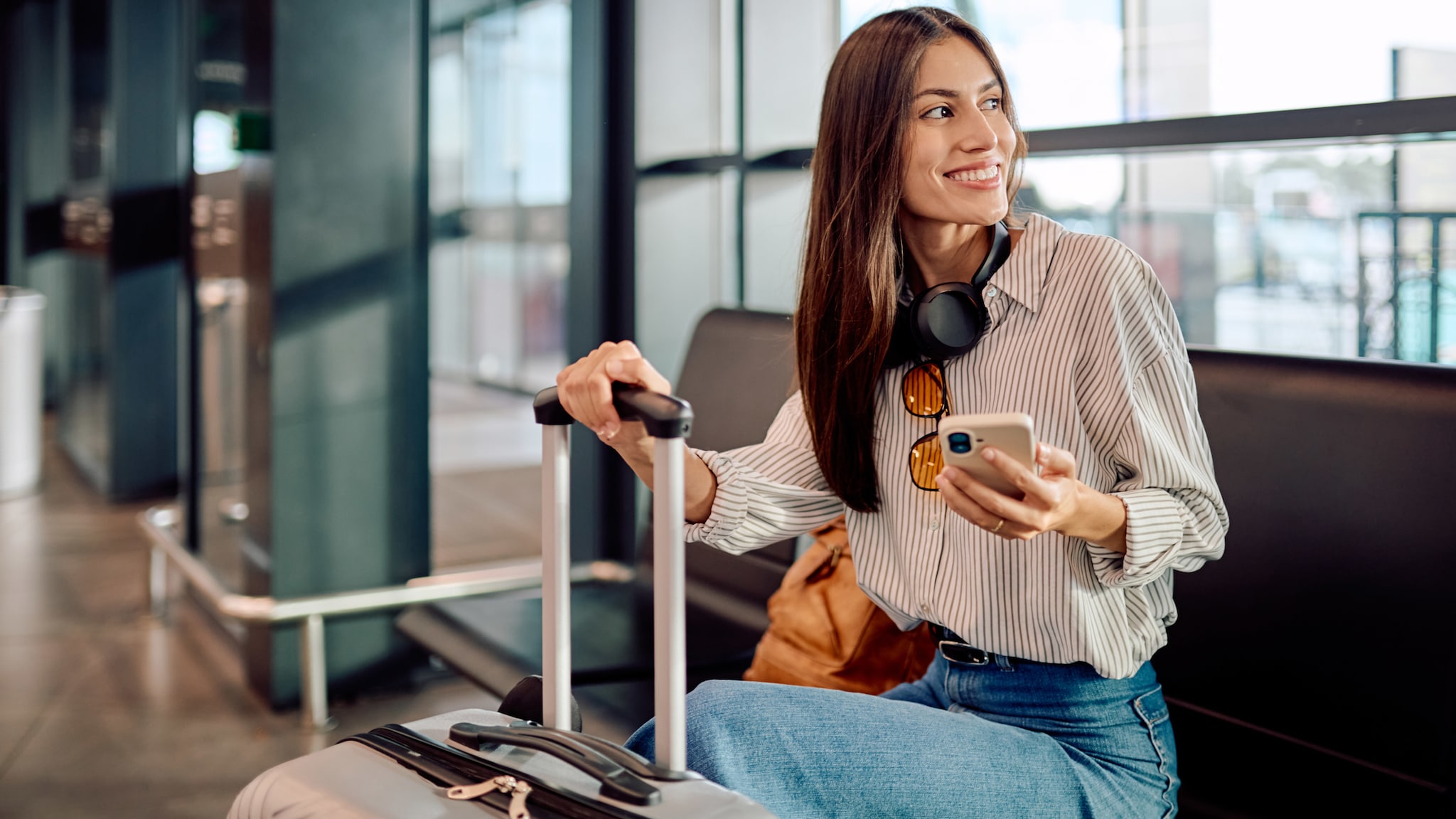 Eine Frau sitzt am Flughafen mit ihrem Koffer und einem Smartphone in der Hand.